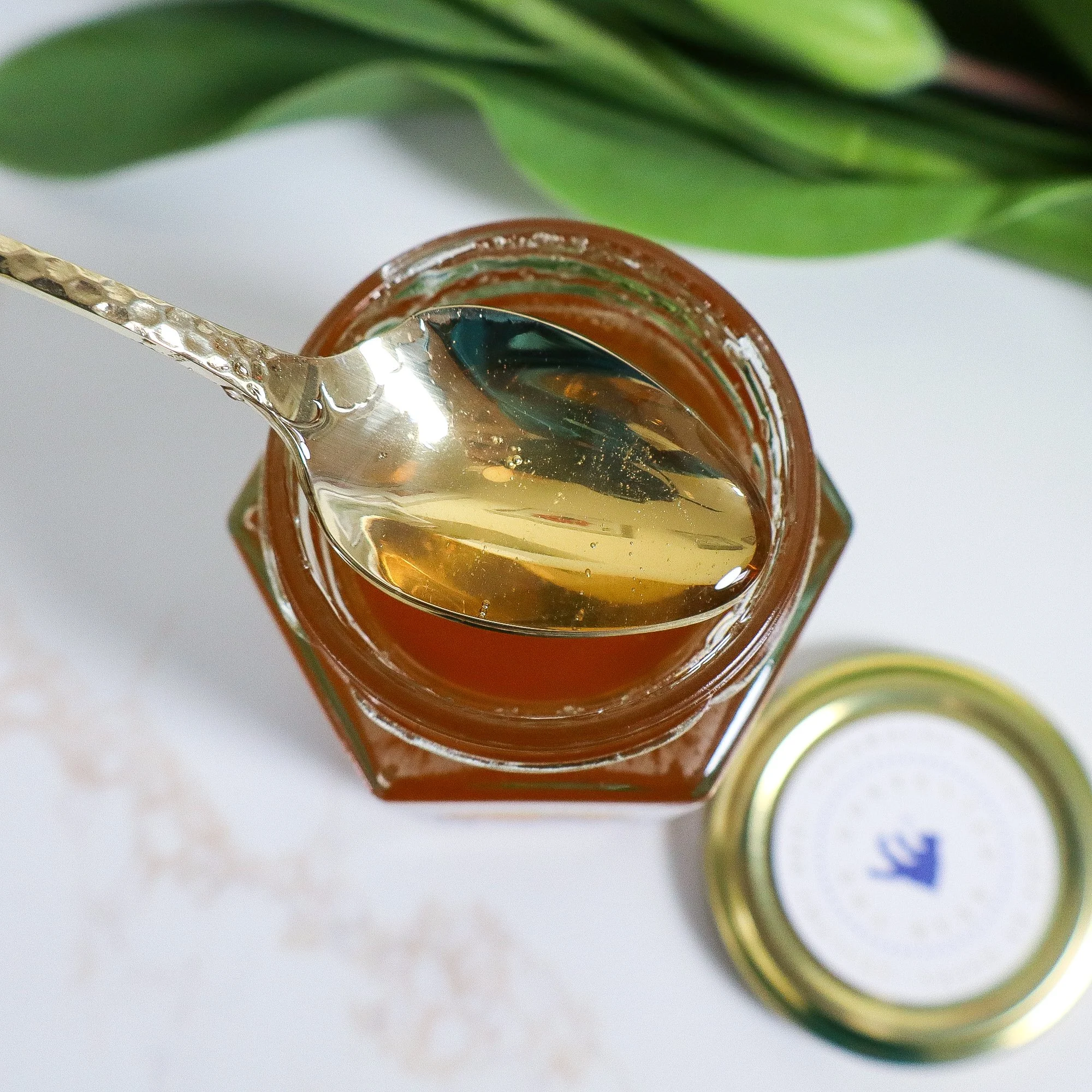 An aerial view of an open hexagonal shaped jar of golden ethical honey with a golden teaspoon brimming with honey above it.