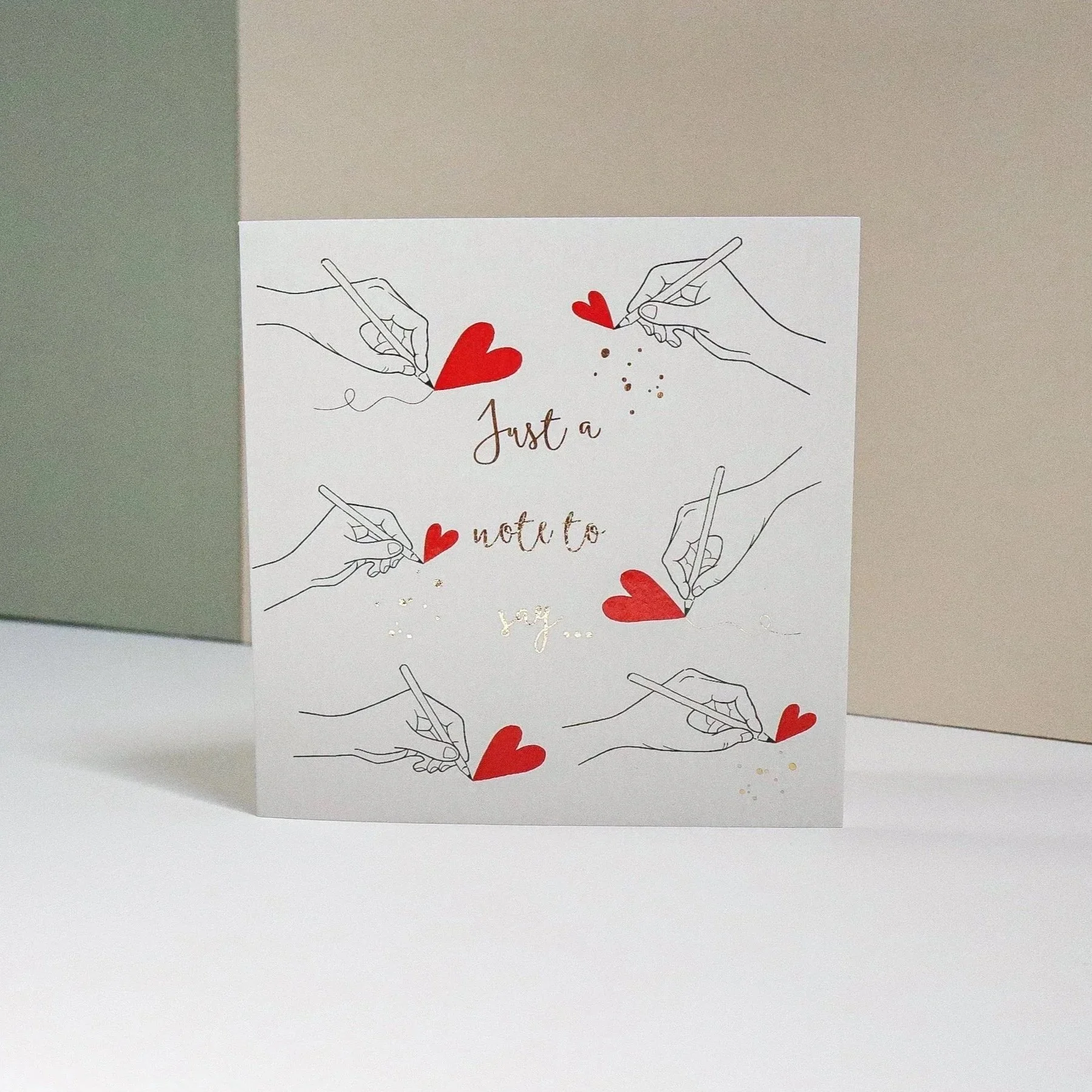 A greeting card standing up with a bicolour pastel colored painted background. The card features hand illustrated hands drawing red hearts and says in gold foil lettering “just a note to say…"