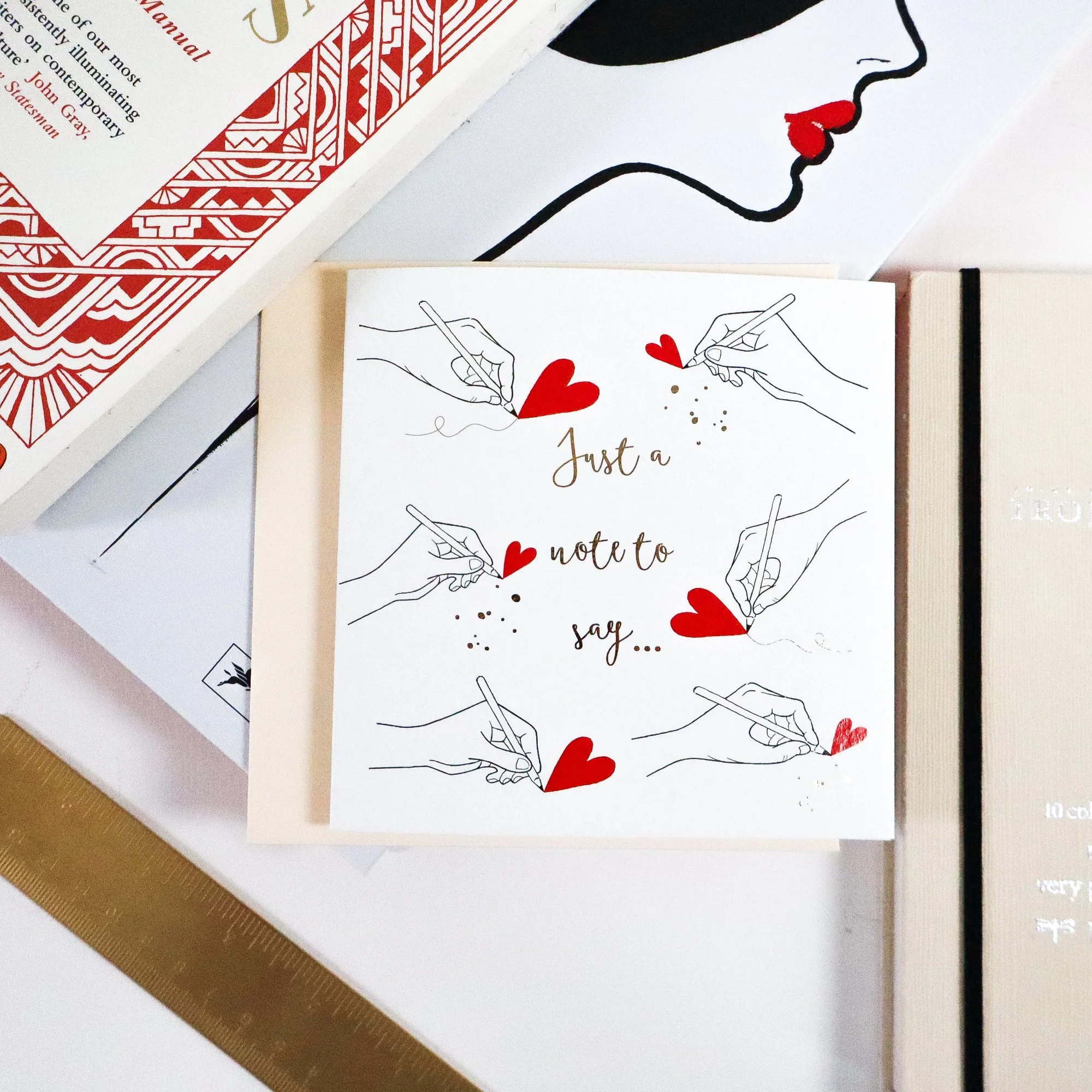 An eco conscious greeting card amongst books and stationery paraphernalia. Everything is cream, red, black or gold.