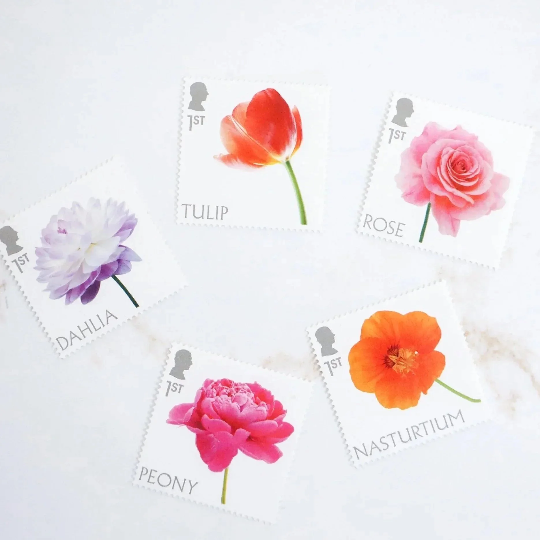 An aerial view of five english stamps each with a different brightly coloured flower on them. They sit on a marble top background.