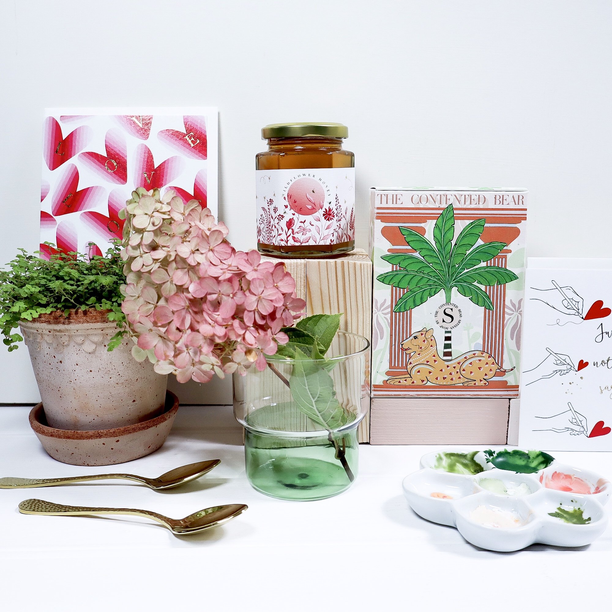 Image of sustainable luxury honey, luxury handmade soap in a lioness illustrated soap box and various terracotta pots and blush coloured flowers in green glasses.