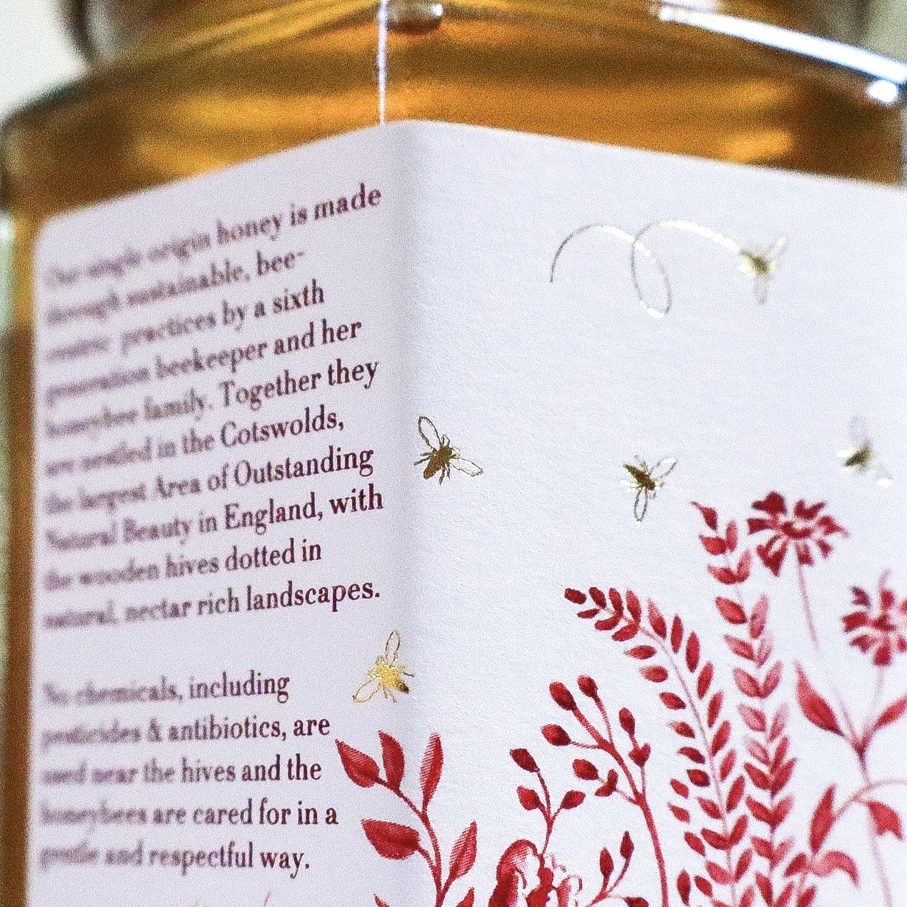 Close up view of a hand illustrated luxury honey label featuring red meadow flowers with gold foil honeybees flying around them.