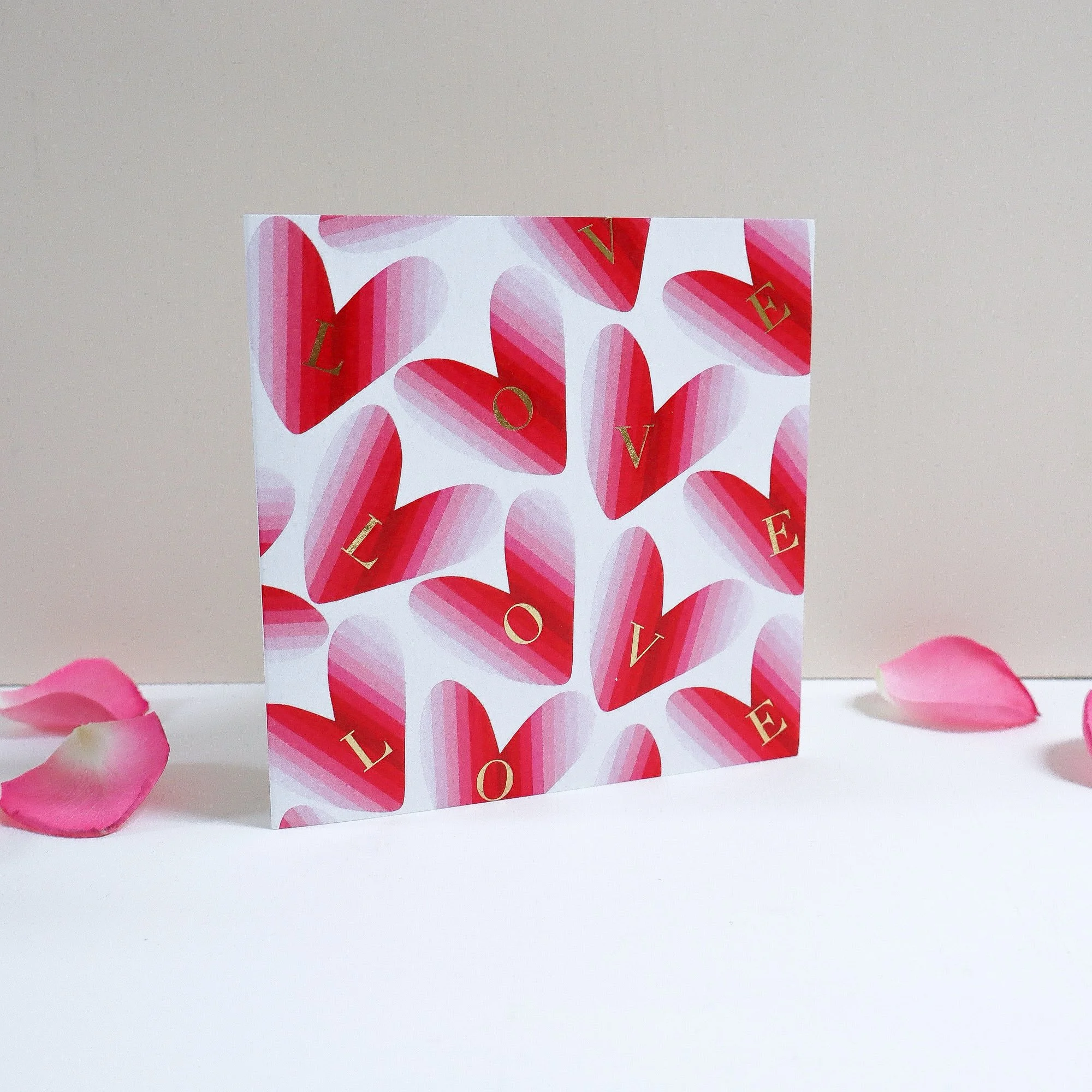 A hand painted greeting card featuring red hearts. The card is standing up and has a few pink rose petals around it with a neutral painted backgropund.