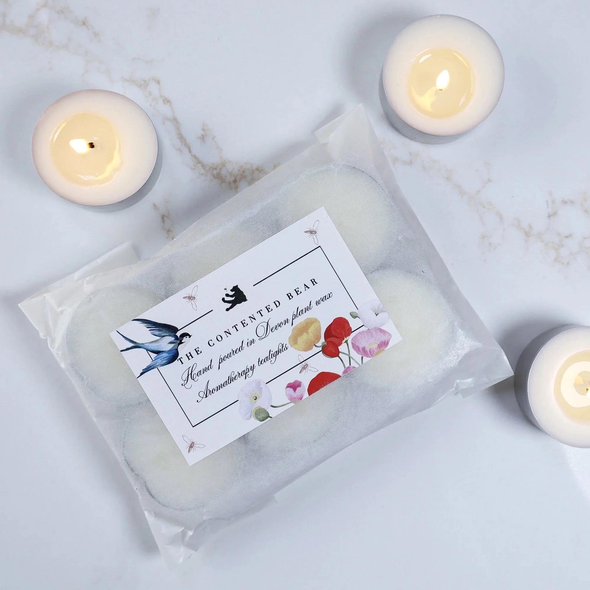 An aerial view of a single packet of artisan, plant wax tea lights with a hand painted label featuring swallows and poppies. The candles are on a white marble background.