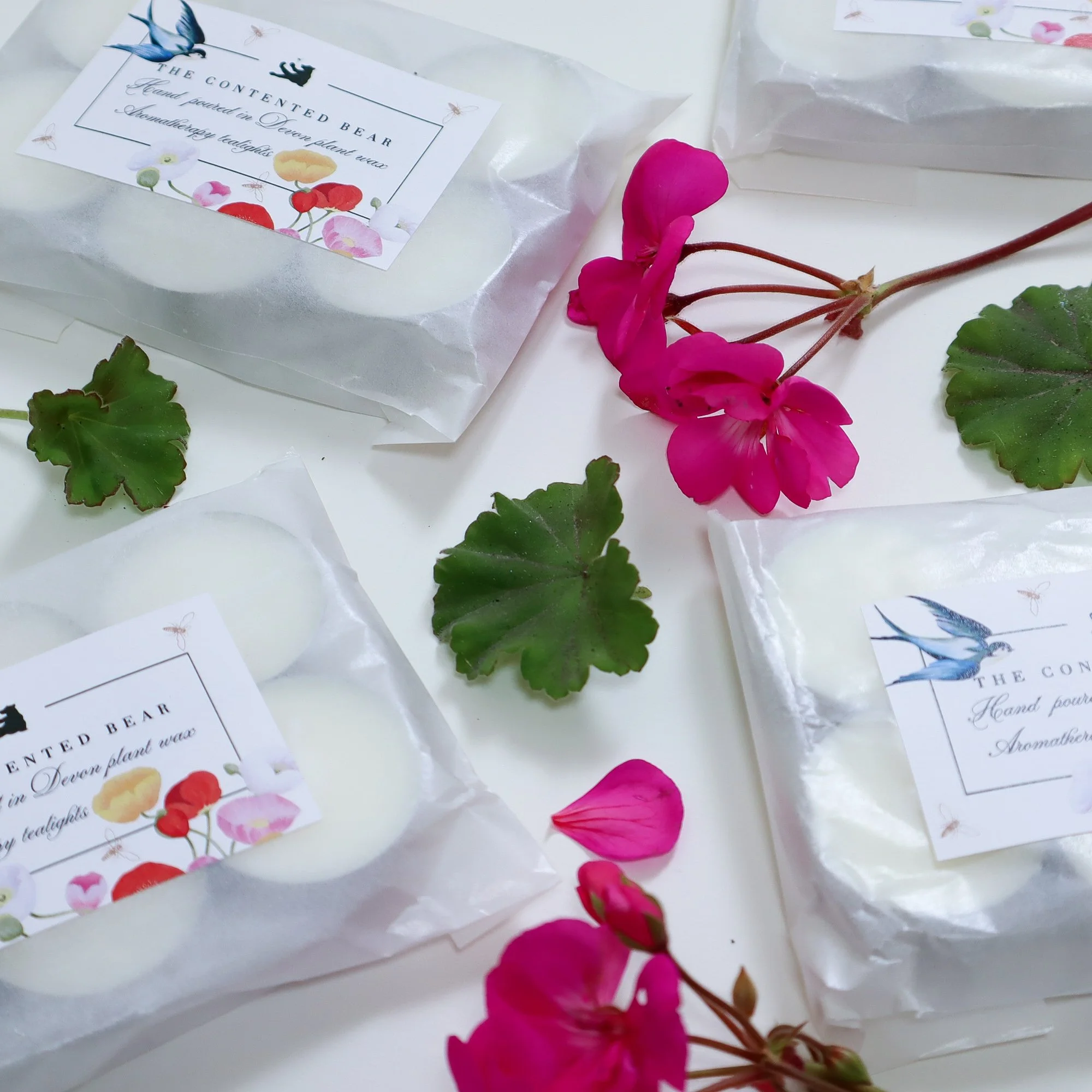 Aerial view of pink rose geranium flowers and leaves with glassine paper packets full of six scented eco tea lights.