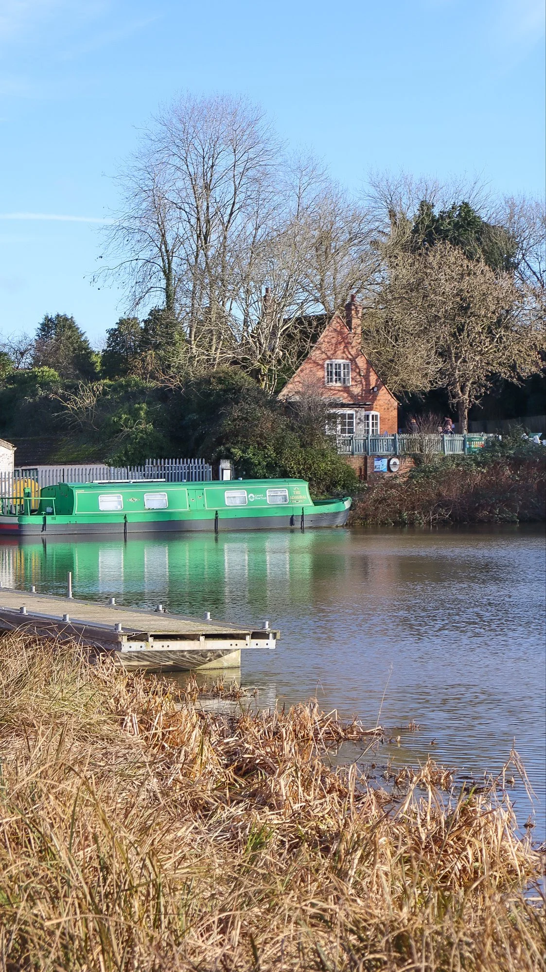 A green canal boat sitting on a small body of water in Caen Hill, Wiltshire, with an old brick built cafe in the background with trees all around and a small jetty to the left.