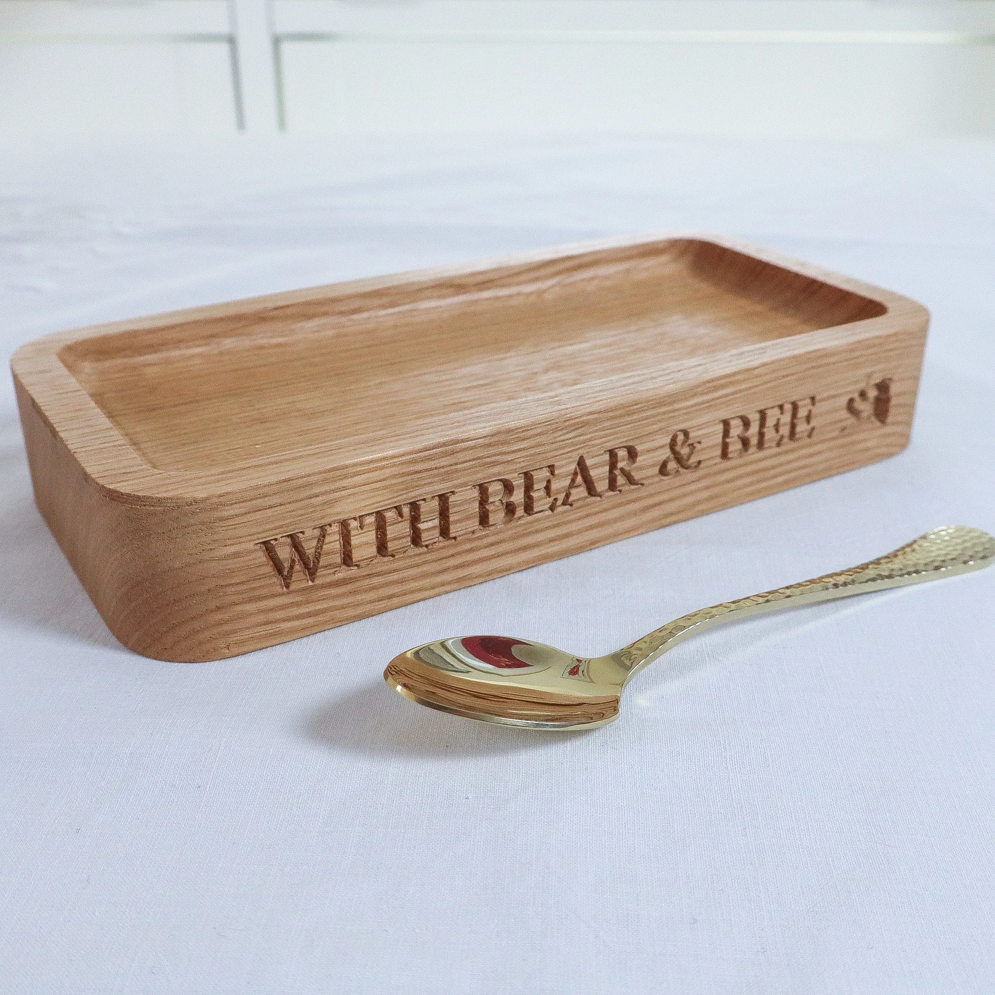 A side view of a hand crafted solid oak tray engraved with text reading ‘with bear and bee’. The tray stands beautifully against a white linen table cloth and a single gold teaspoon.