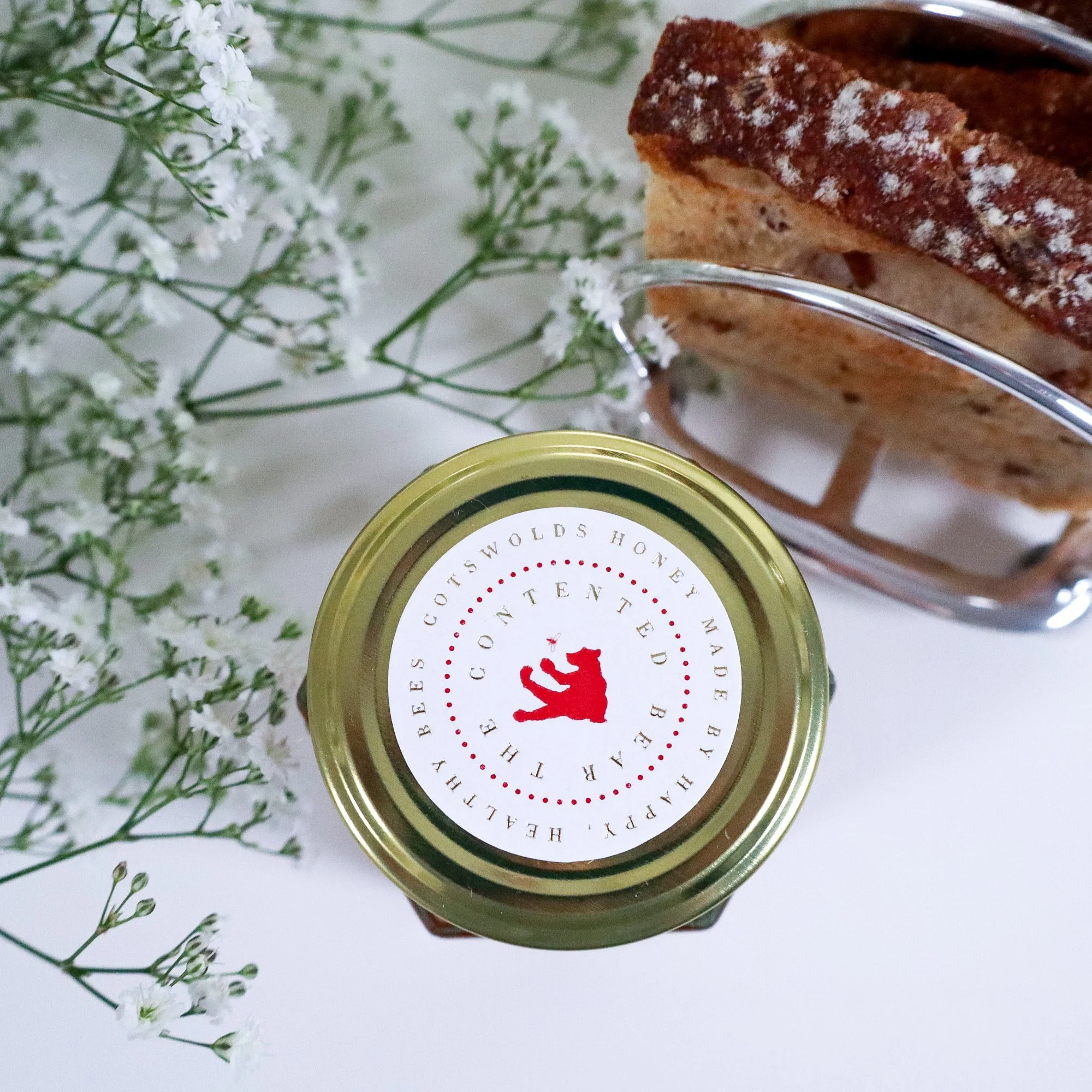 Aerial view of luxury, sustainable honey with delicate white flowers to the top left and top right is a silver toast rack with brown artisan toast.