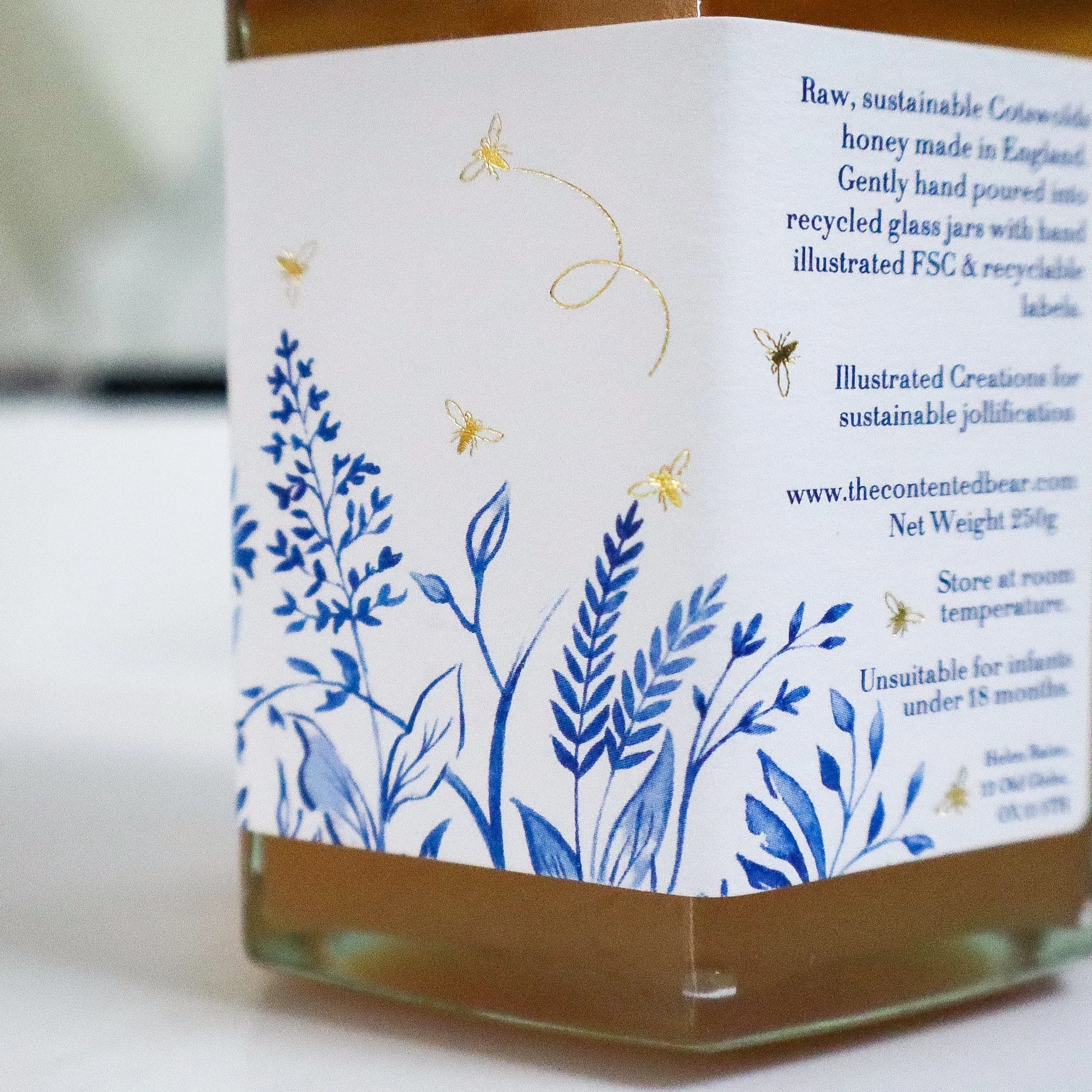 Close up side view of the hand illustrated label of a jar of sustainable honey. The luxury label features blue wildflowers and gold foiled honeybees.