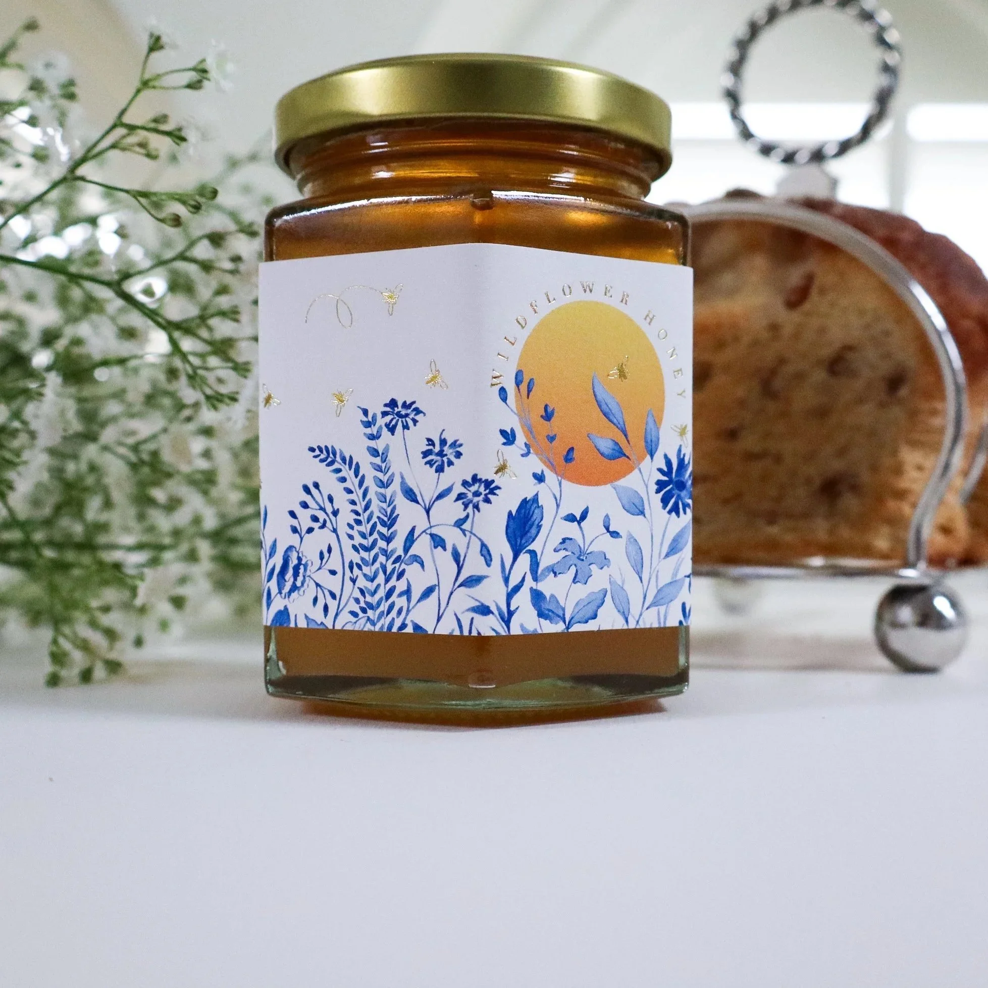 A side view of a beautiful jar of luxury, ethical honey with a blue, white and yellow hand illustrated label and a silver service background.