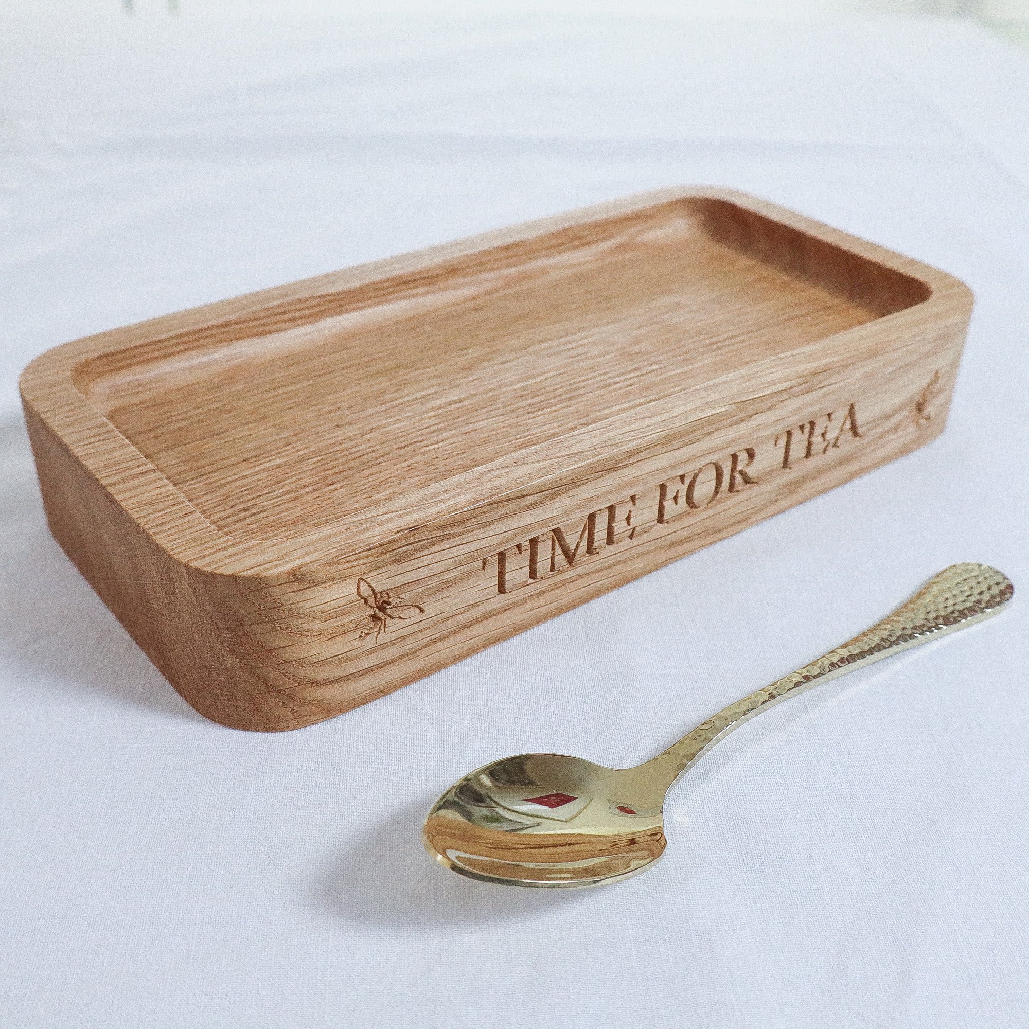 A side view of a solid oak tray engraved with the text ‘time for tea’. The beautiful tray stands on a white linen table cloth with a single gold teaspoon.