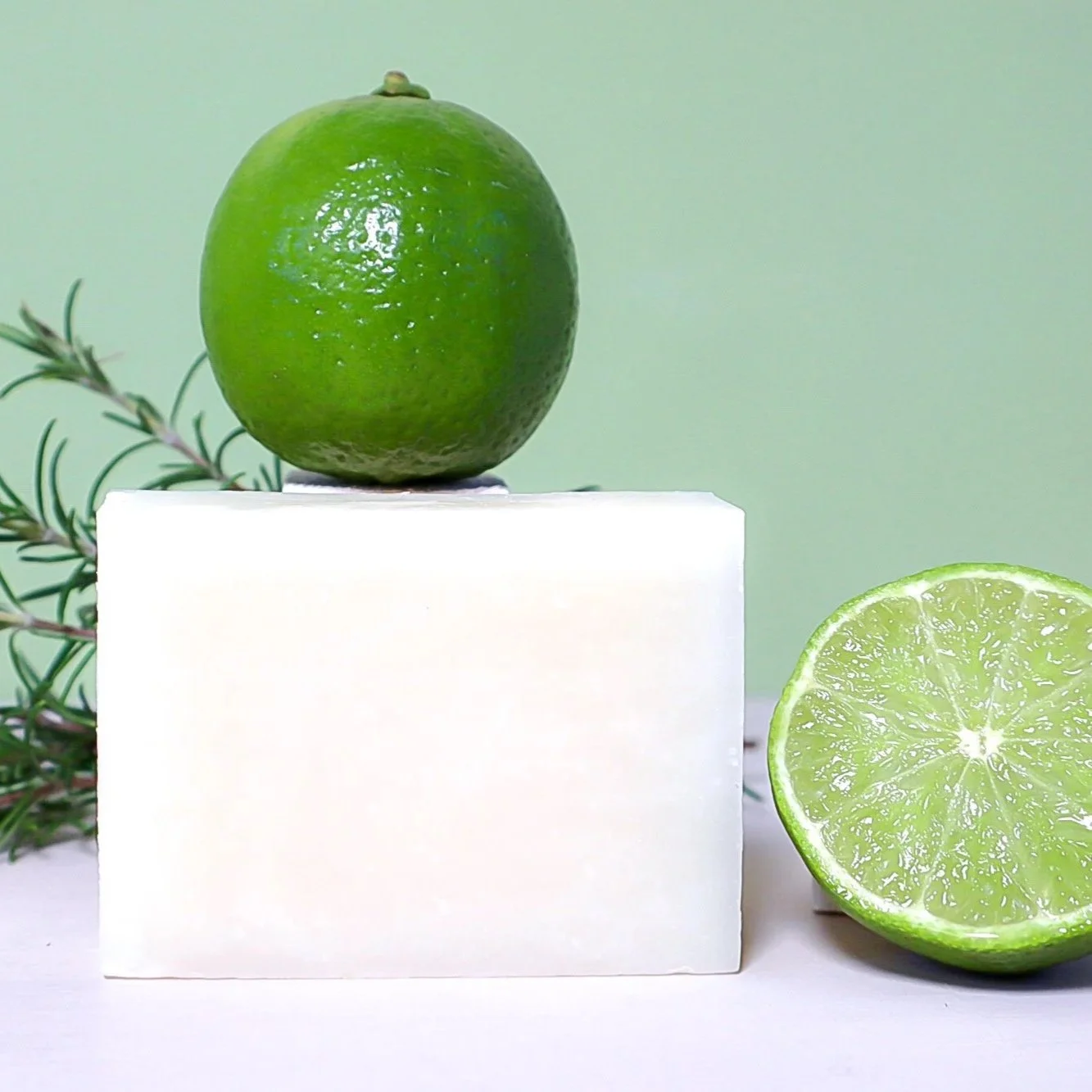 A hand crafted bar of artisan soap featuring the ingredients of fresh rosemary on the left and a fresh green lime above the soap and haf a fresh lime on the right of the soap.