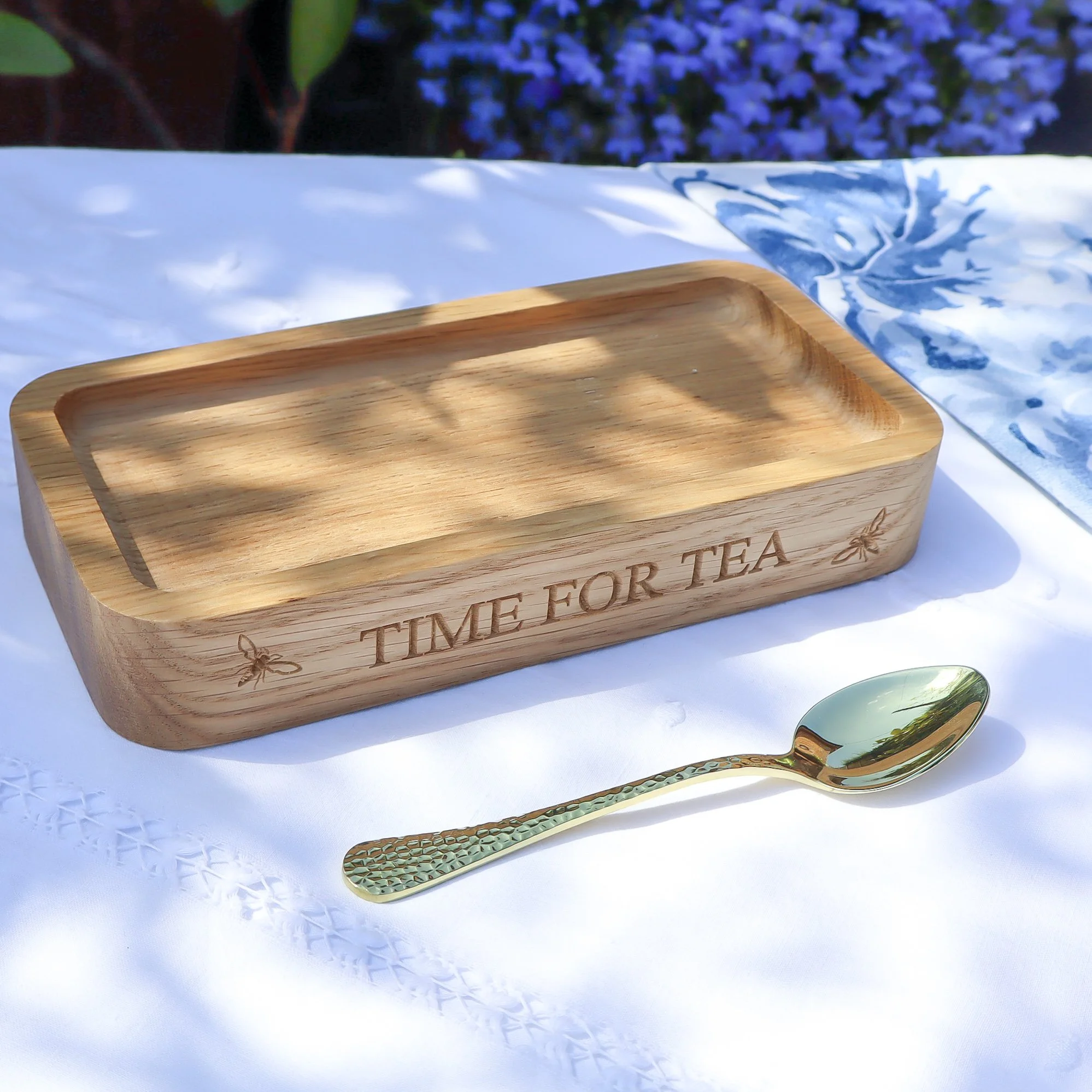 Side on view of an engraved sustainable oak tea time tray. It sits on a white linen table cloth with a blue and white napkin and a golden teaspoon. The backdrop is blue lobelia and green leaves.
