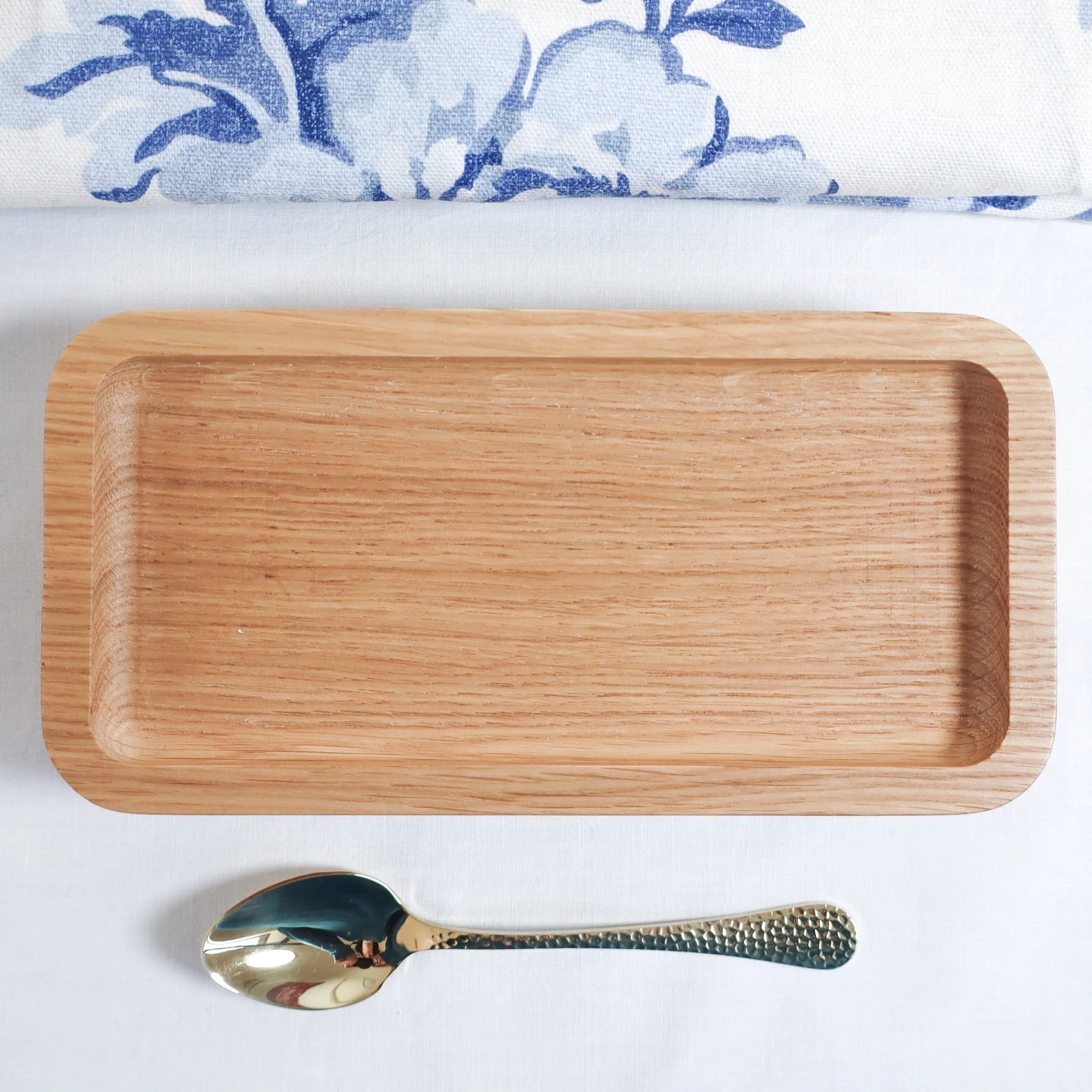 An aerial photo of solid oak tray hand made in britain with a blue and white linen knapkin above it and a single gold teaspoon below it. They all sit on a white linen table cloth.