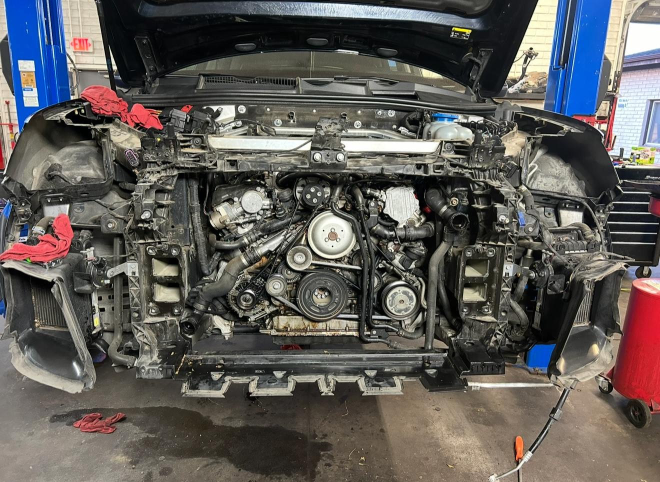 Front view of a car with the hood open, showing the engine and various mechanical components, in a garage or workshop setting.