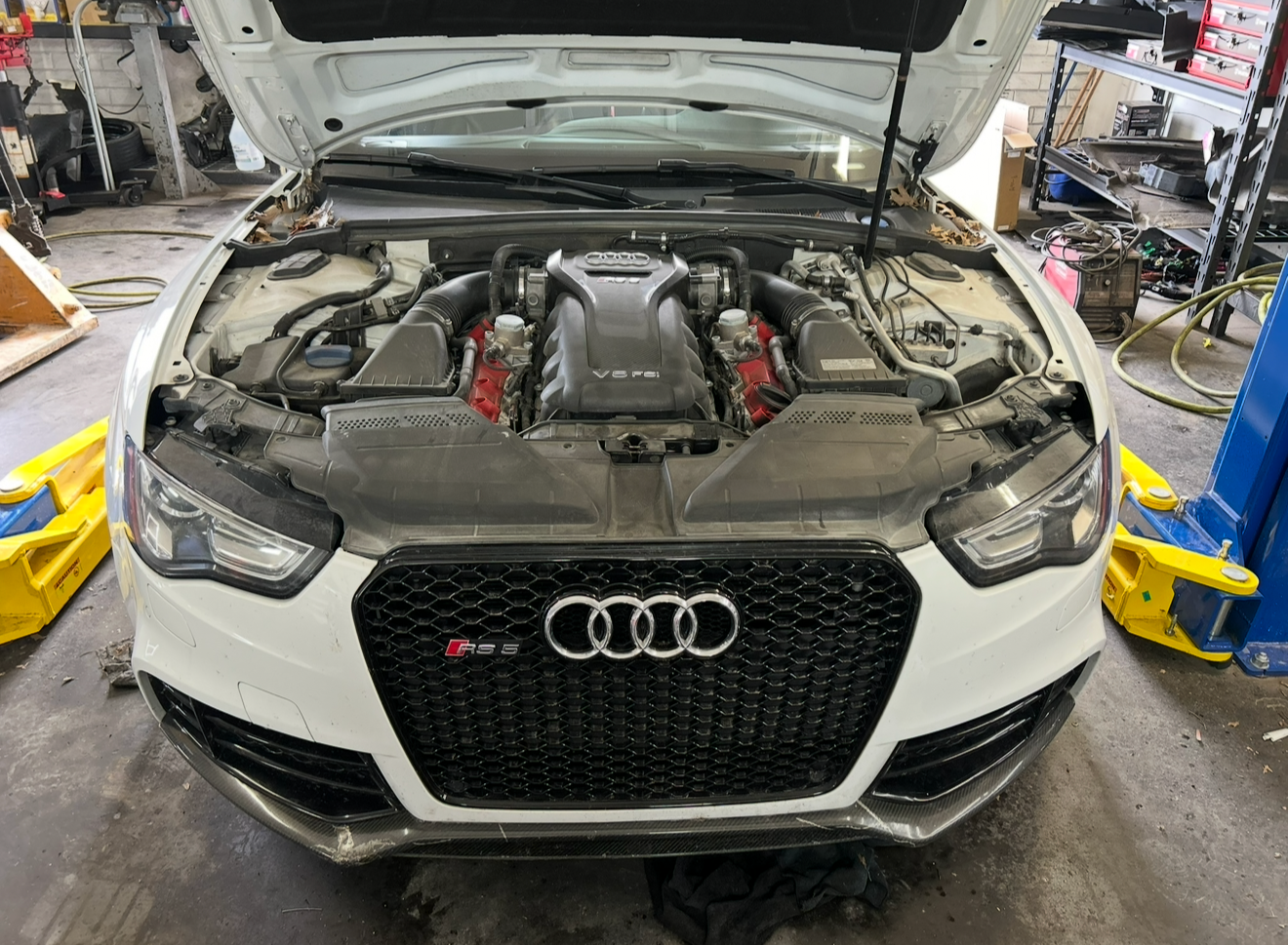 Close-up front view of a white Audi RS 5 with its hood open, showing the engine bay in a garage or workshop setting.