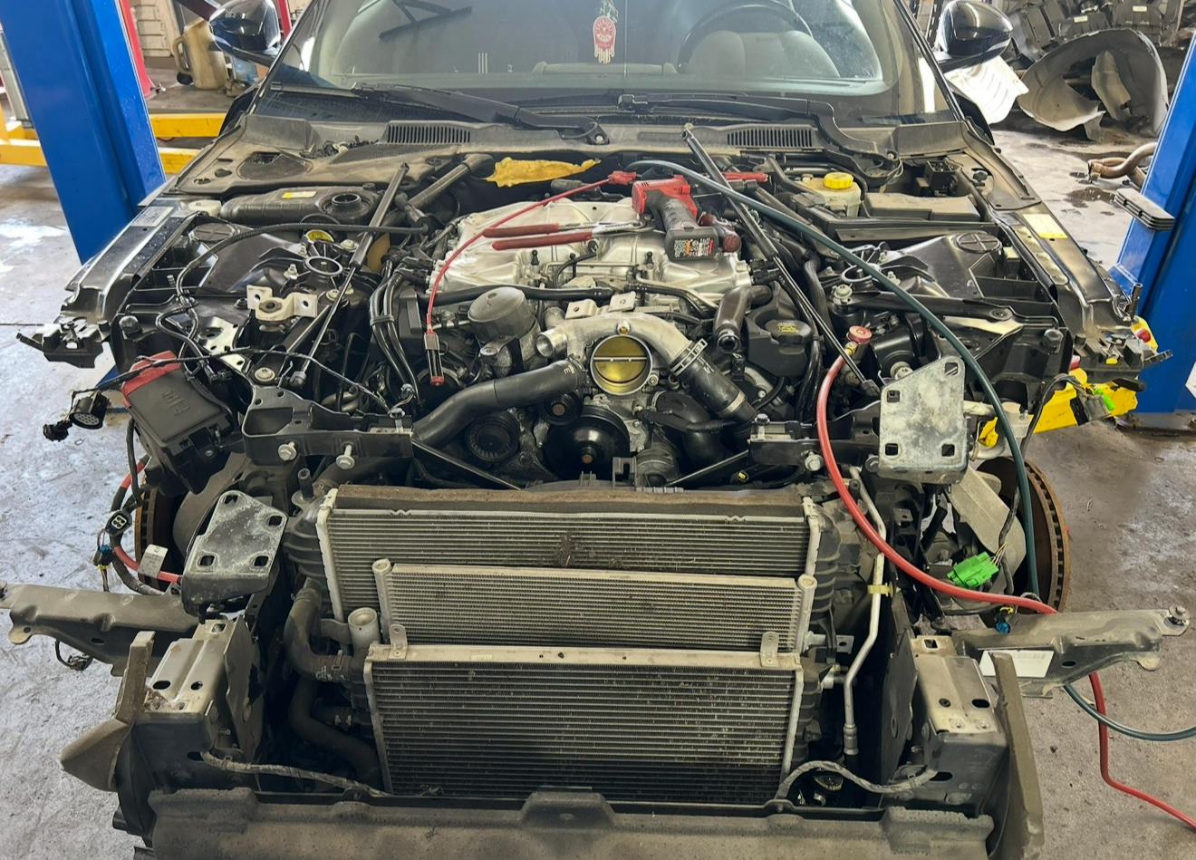 Front view of a car with its hood open, showing the engine and radiator, in a repair shop with tools and other vehicle parts in the background.
