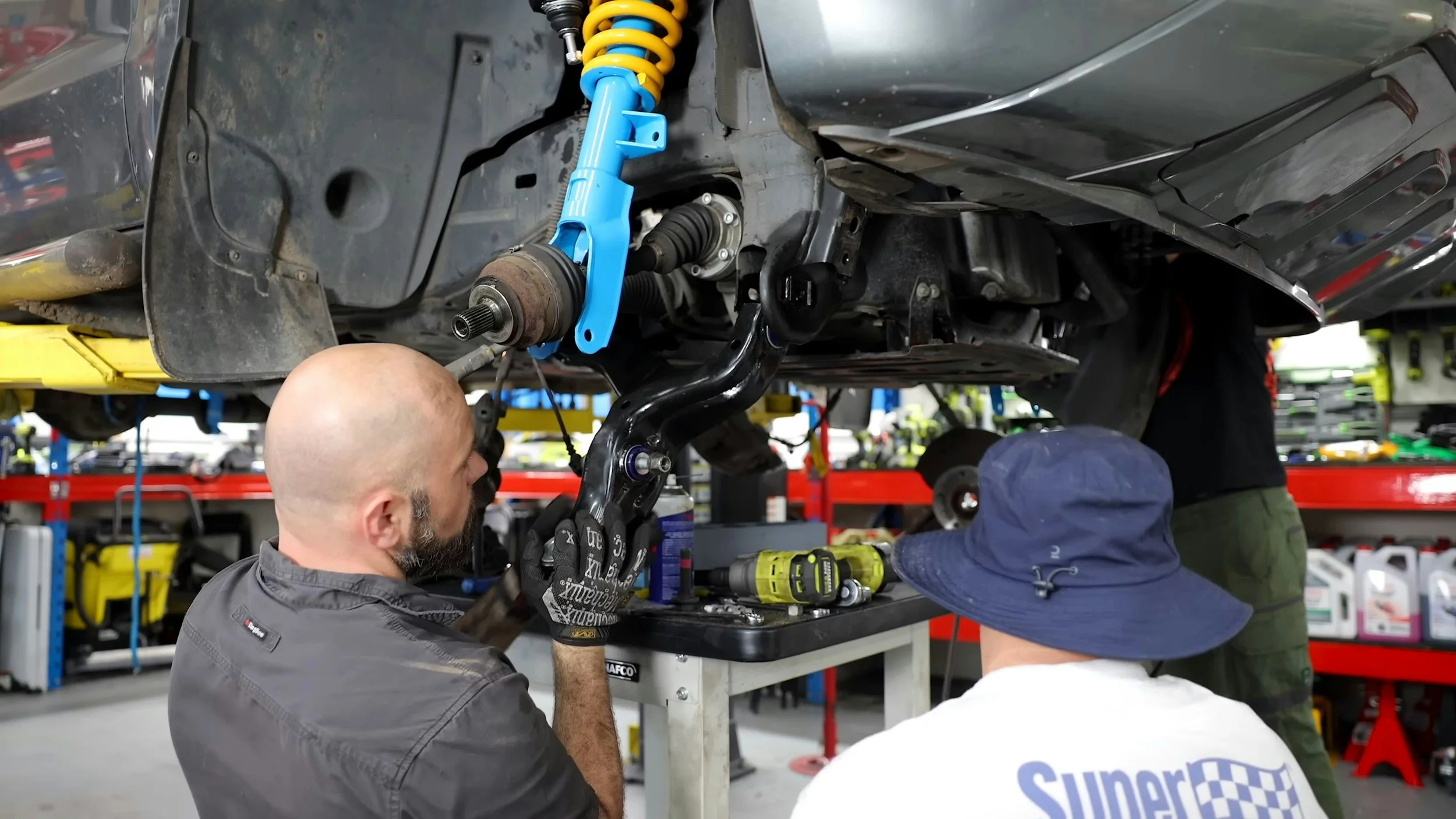 Two mechanics working on a car suspension system in an auto repair shop, with tools on a workbench and shelves of automotive supplies in the background.