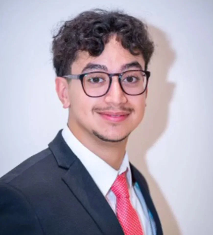 A young man with curly dark hair, glasses, and a light mustache, wearing a black suit, white shirt, and pink tie, smiling at the camera against a plain light background.