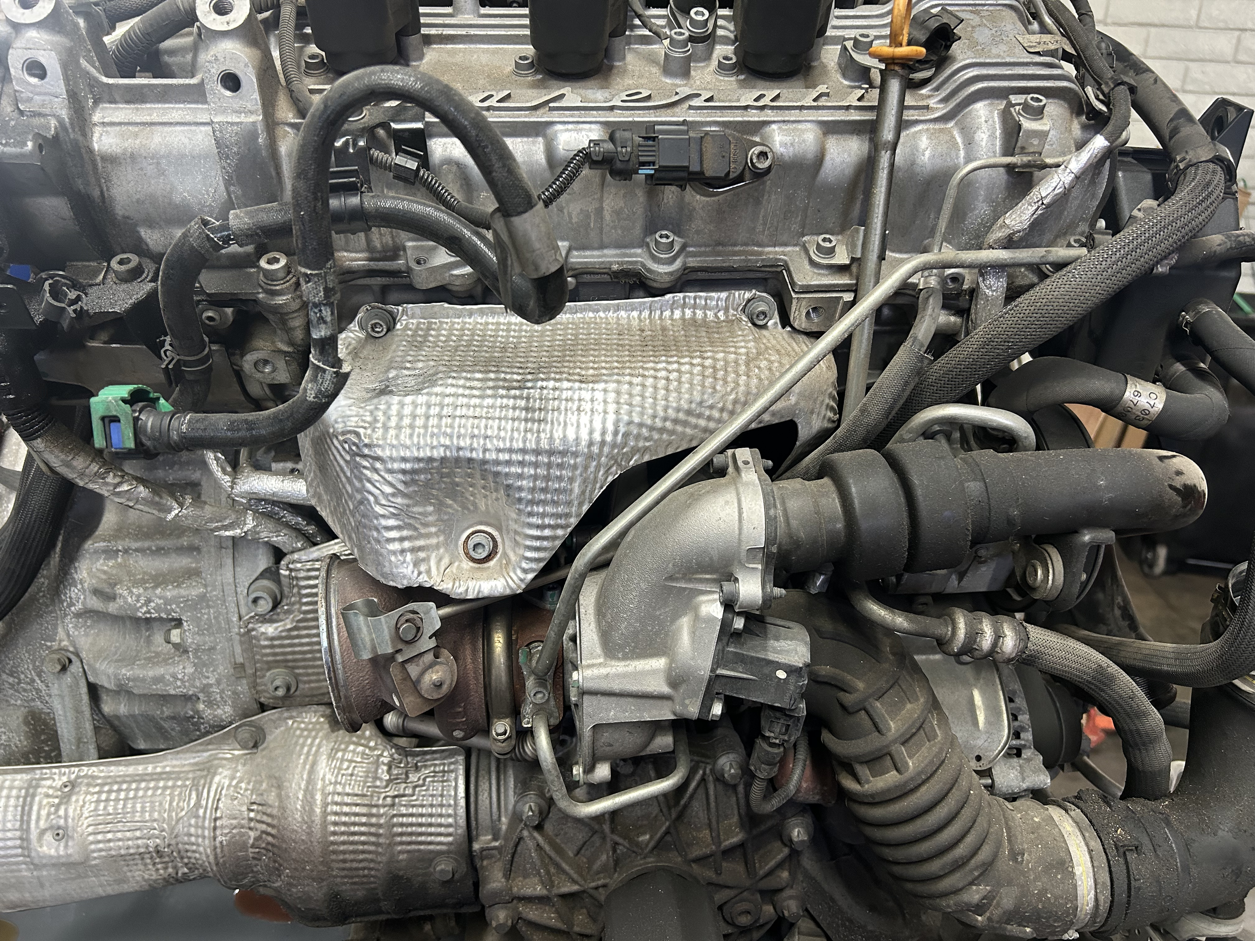 Close-up of a car engine with various hoses, pipes, and metallic components, some covered with heat shields, in a workshop setting.