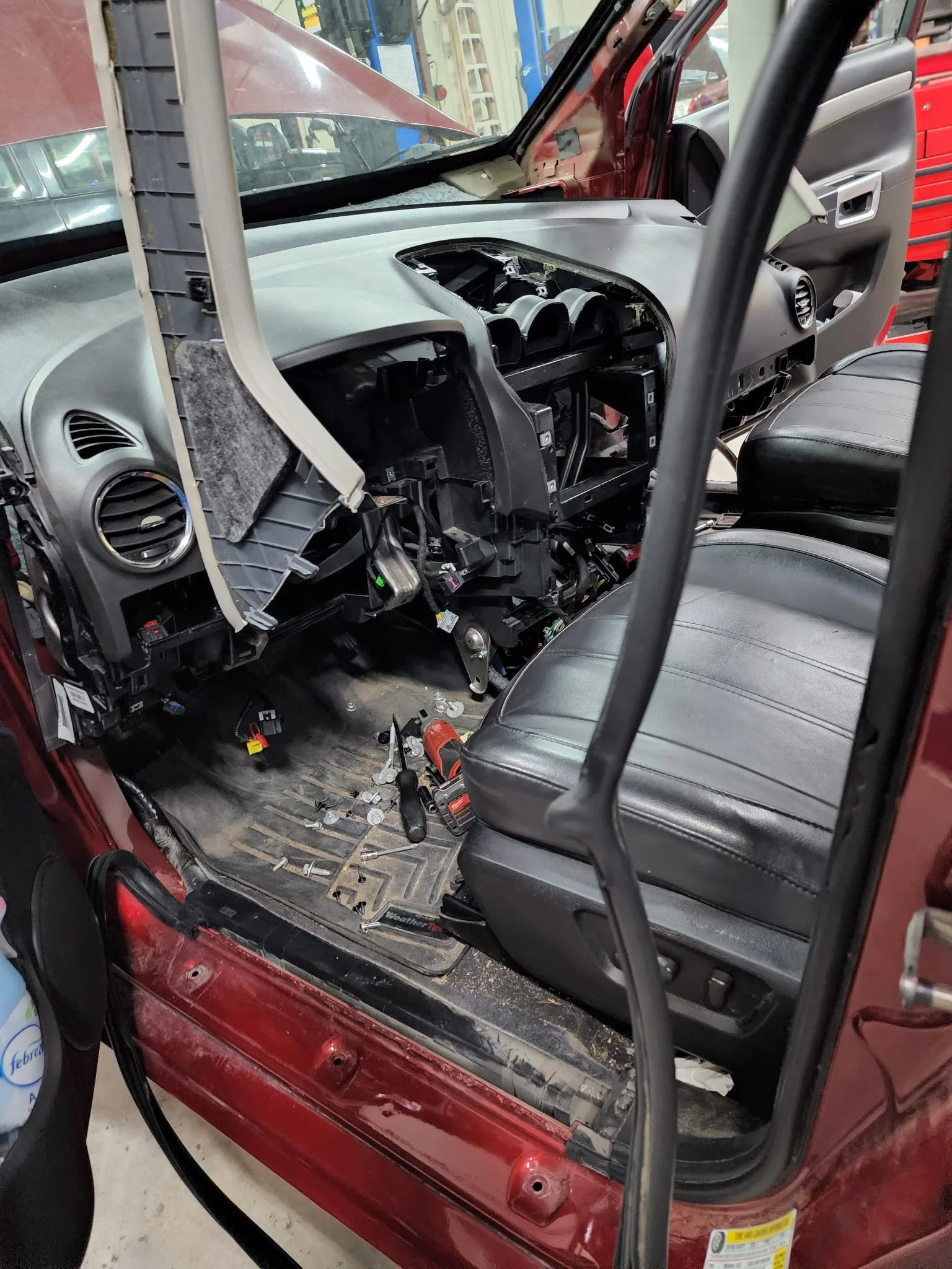 The interior of a vehicle undergoing repairs, with the dashboard partially disassembled, exposing internal components. Tools and parts are scattered on the floor near the driver's seat.
