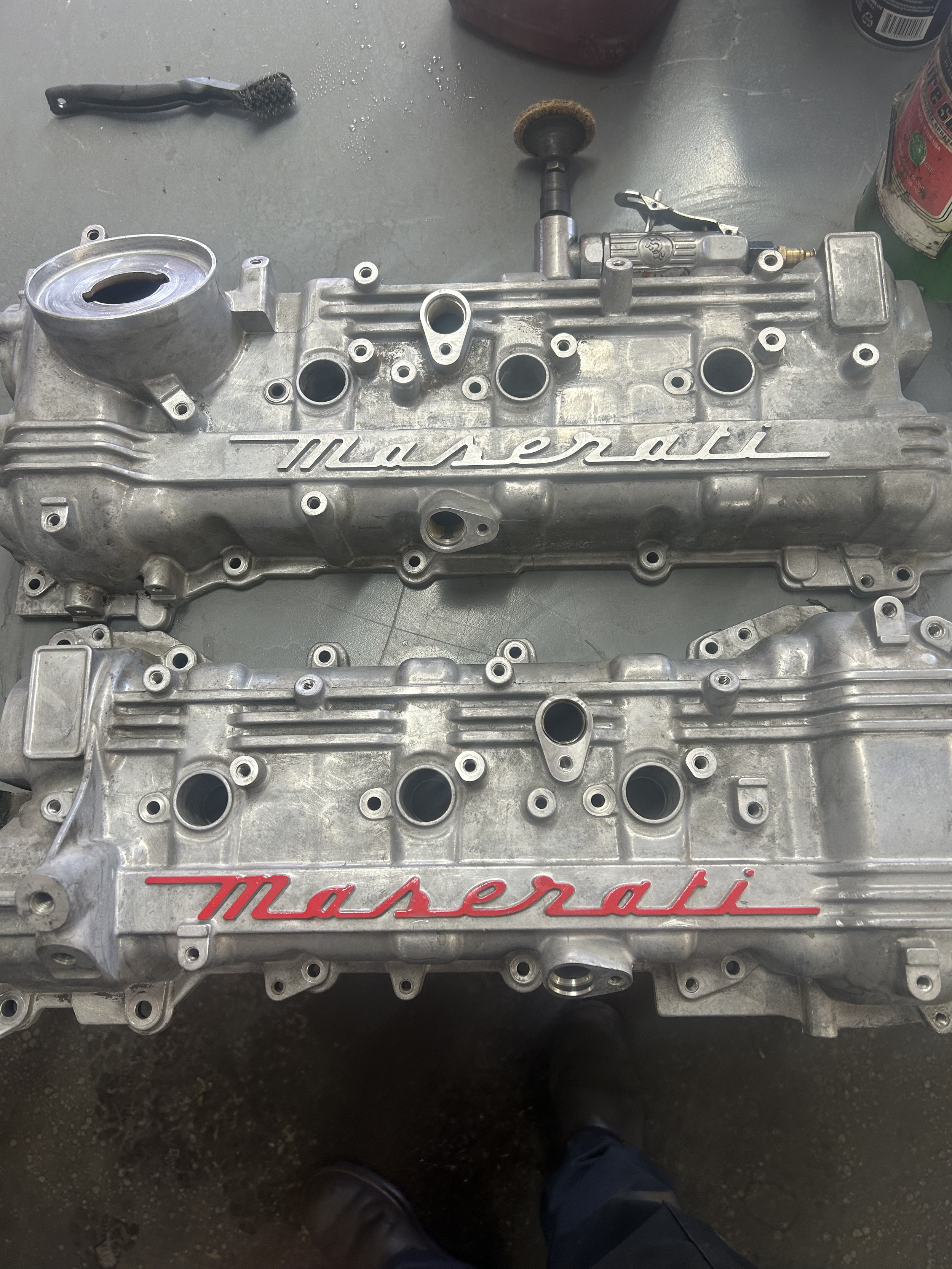 Two silver Maserati engine valve covers on a gray work surface with tools and spray cans around.