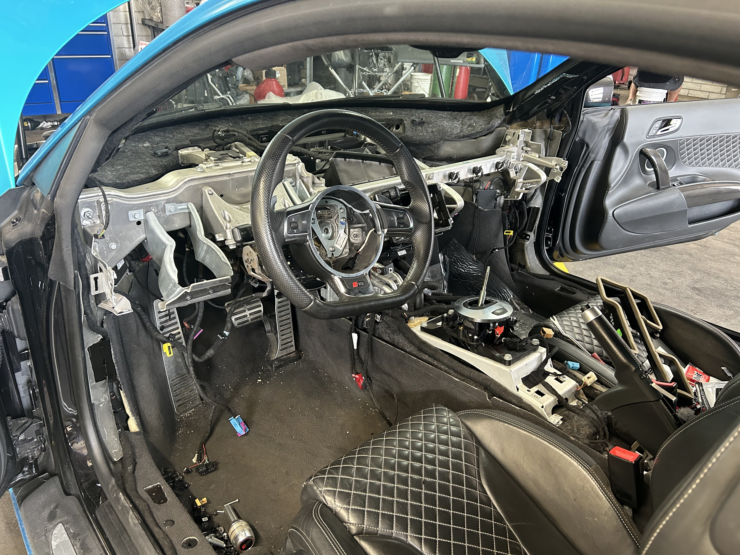Interior of a partially disassembled race car showing the dashboard, steering wheel, pedals, and seats, with exposed wiring and metal components.