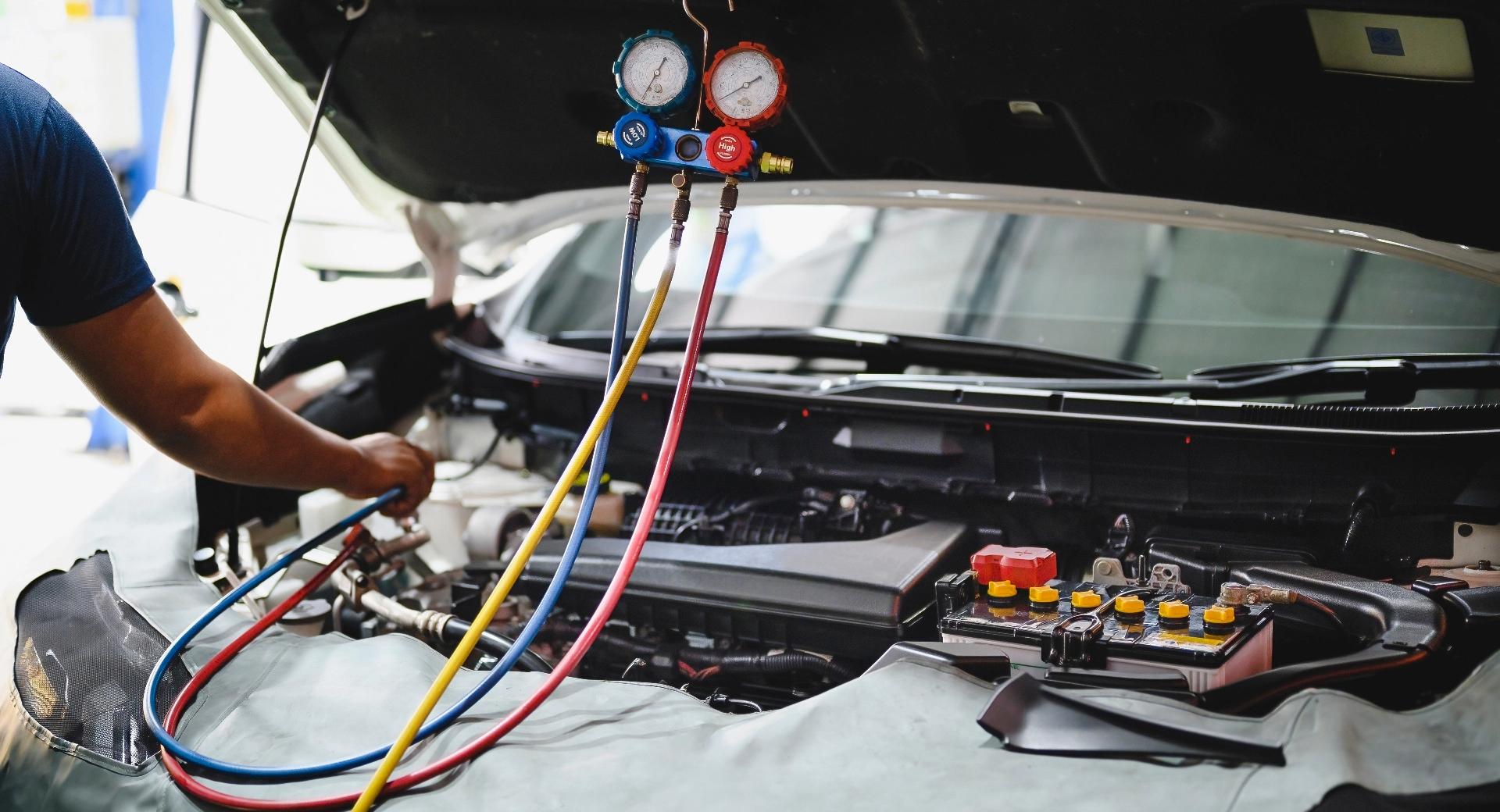 Automotive technician using gauges and hoses to perform air conditioning service on a car with the hood open.