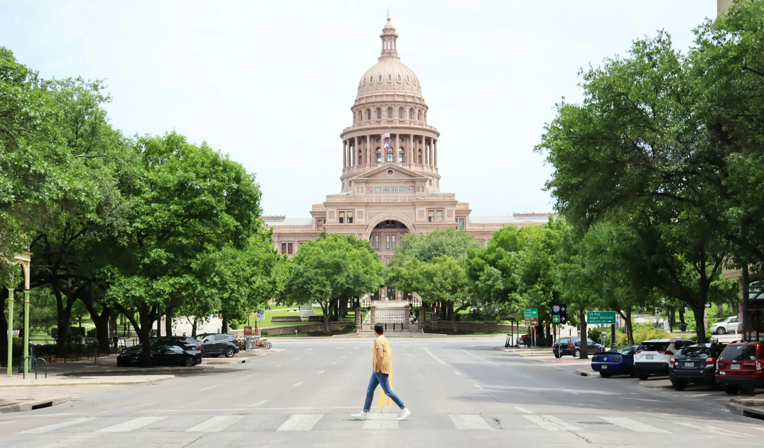 A person walking across a crosswalk towards the Texas State Capitol building, surrounded by green trees and parked cars.