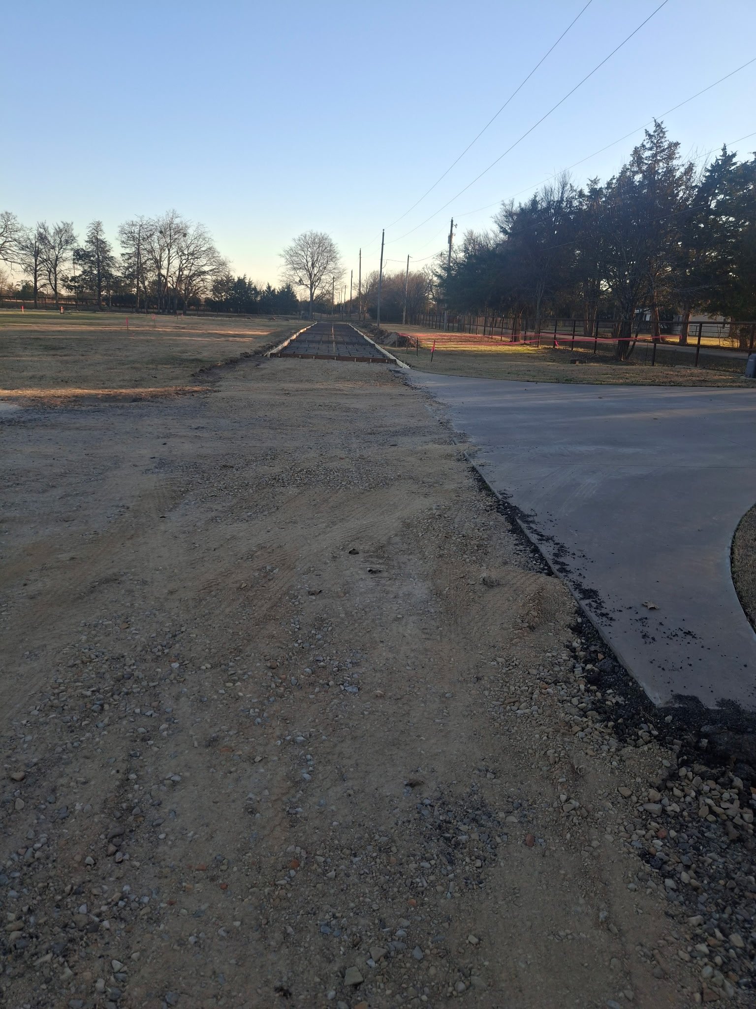 A construction site where a new sidewalk is being built on the side of a road, with gravel and concrete visible, trees and utility poles in the background, and a clear blue sky overhead.