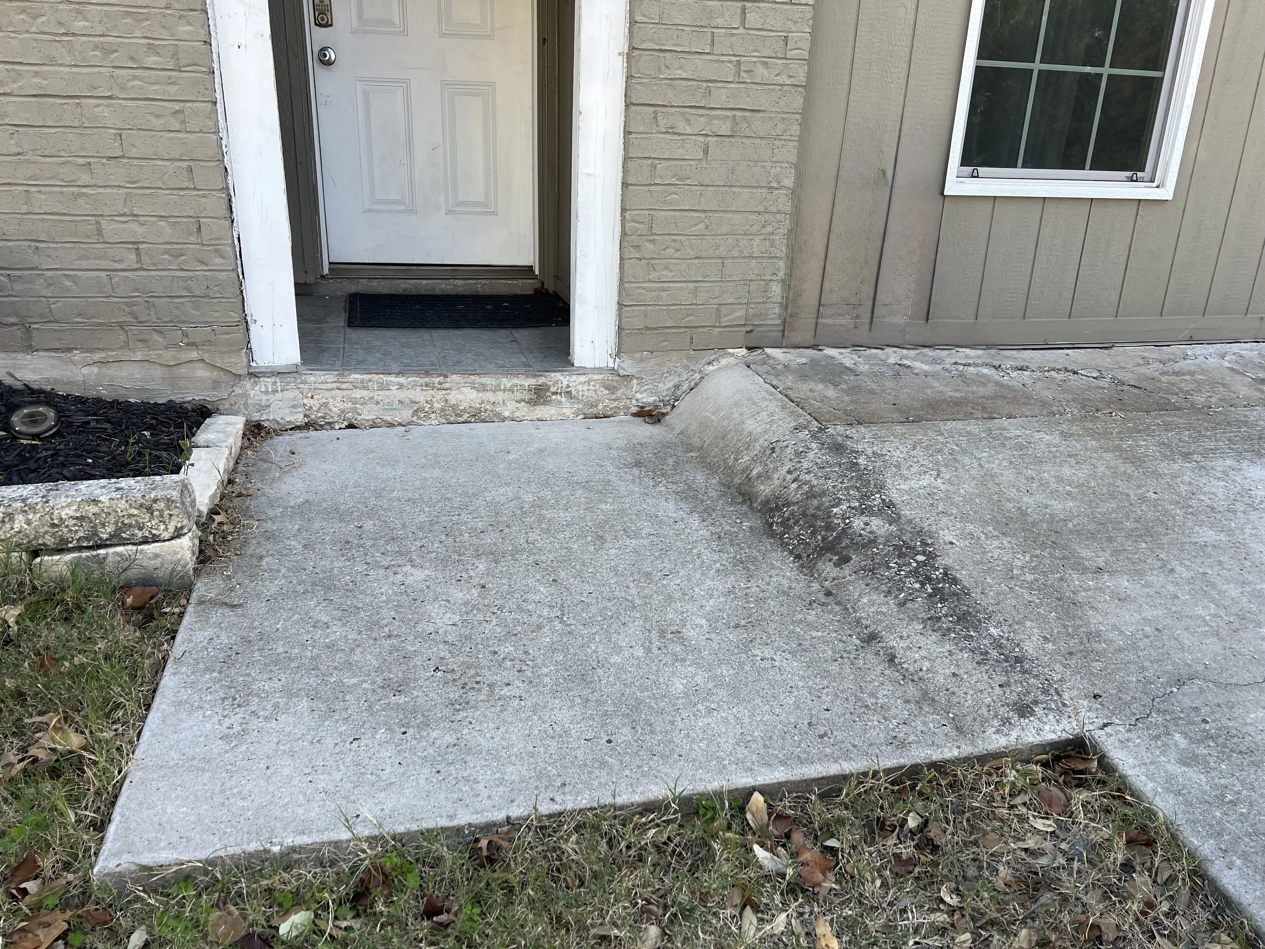 Concrete front step and sidewalk in front of an apartment entrance, with a small landscaped area on the left and a window on the right.