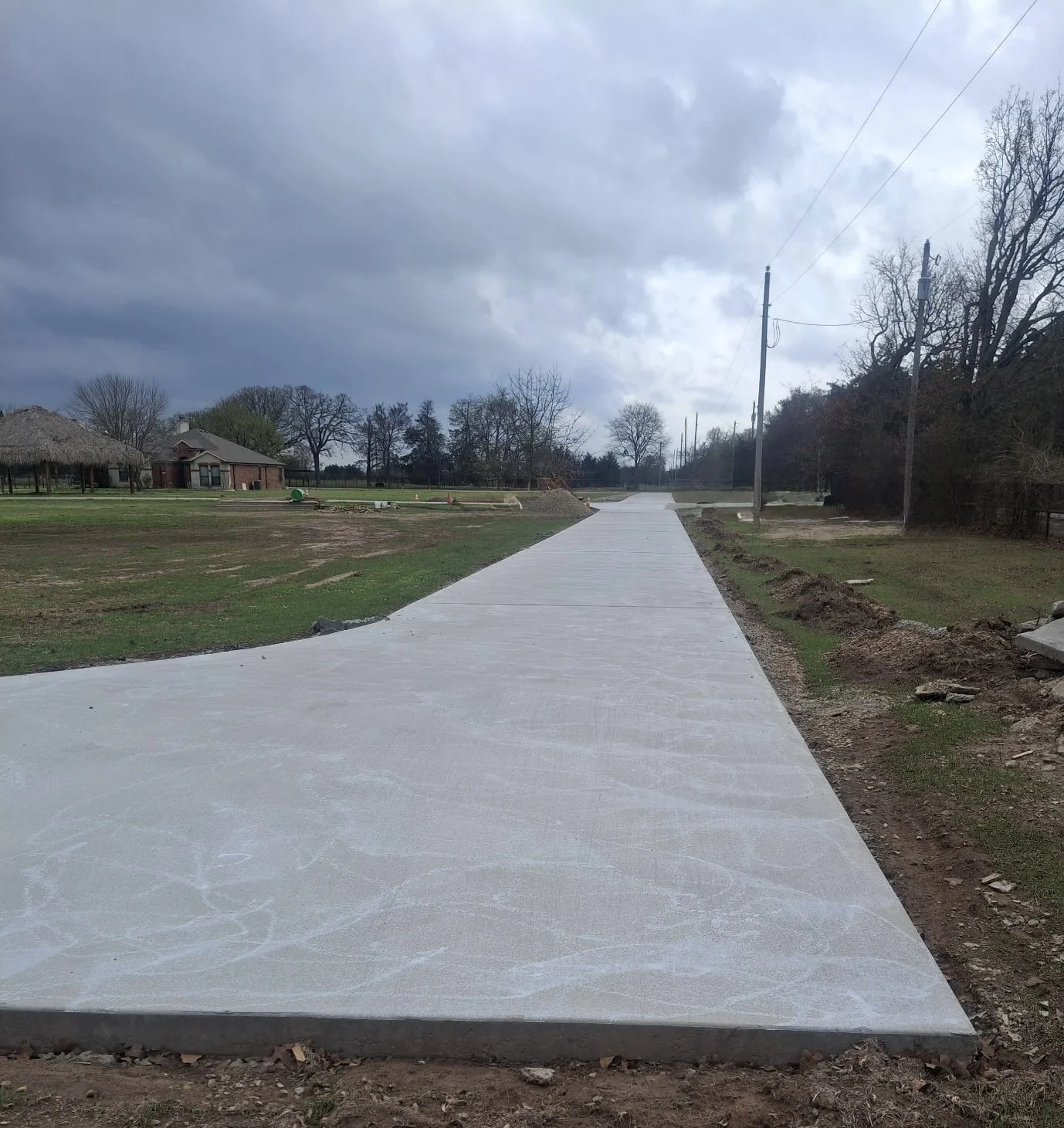 Newly poured concrete sidewalk in a rural area under cloudy sky with utility poles and trees in the background.