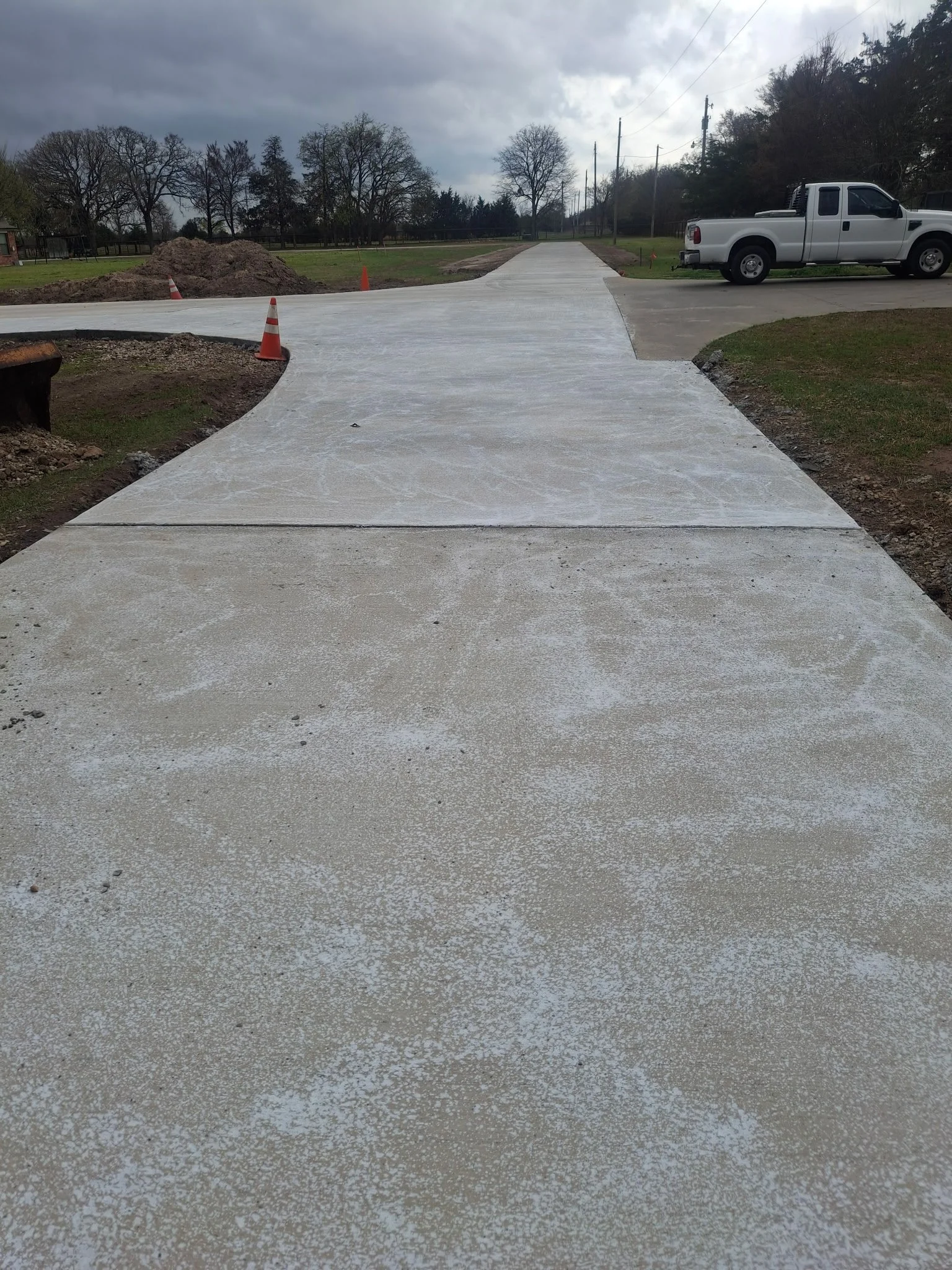 Newly poured concrete sidewalk with construction cones on the side, outdoors under a cloudy sky, with an area of grass, trees, and parked vehicles visible in the background.