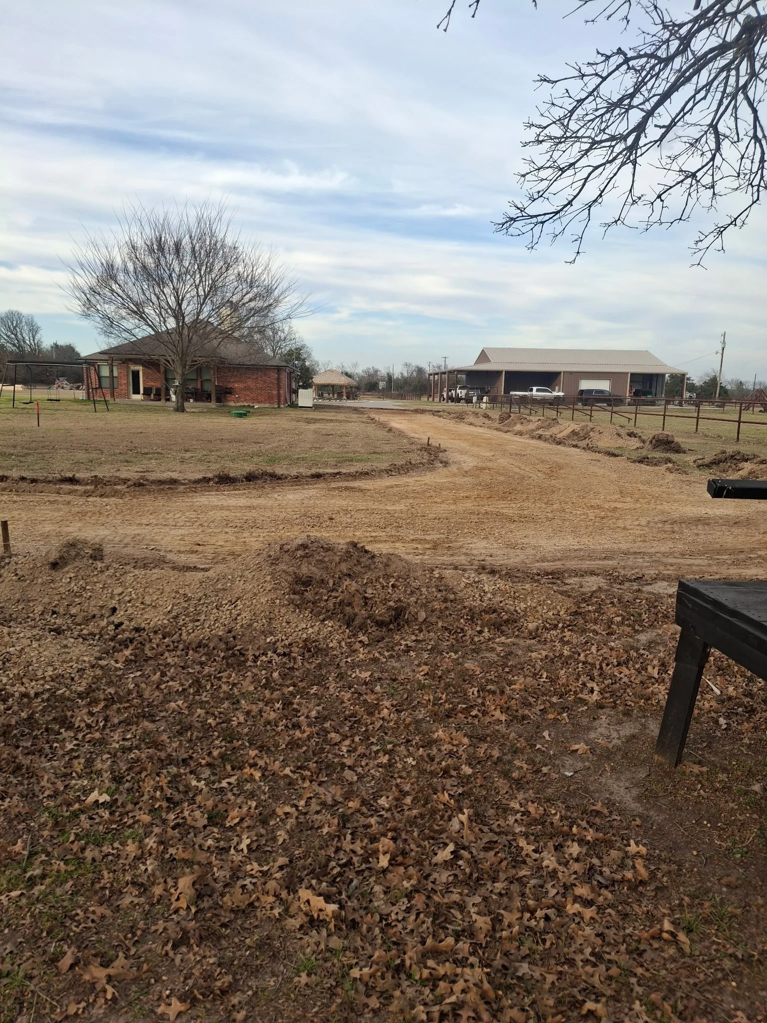 A rural scene with a dirt road, leafless trees, a brick house on the left, a large barn on the right, and cloudy sky overhead.