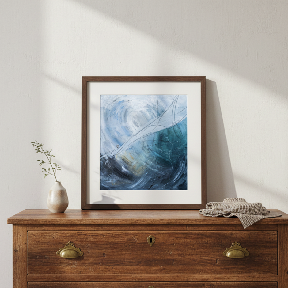 A framed abstract painting with blue, black, and white swirls, leaning against a white wall. To the left, a small vase with a green plant. To the right, a folded cloth. The furniture is a wooden dresser with brass handles.