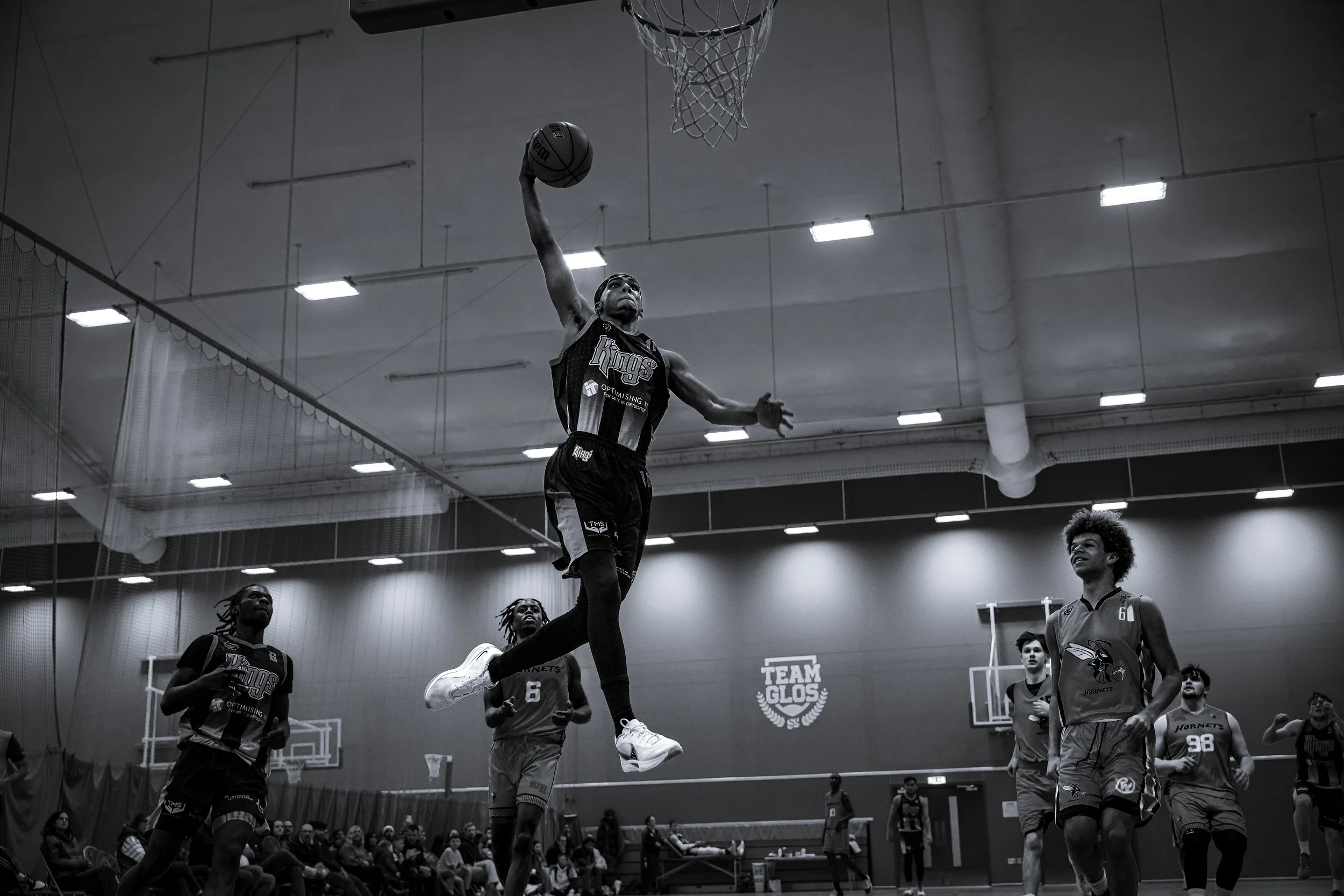 A basketball player is leaping towards the hoop for a dunk during a game, with other players and spectators in the background.