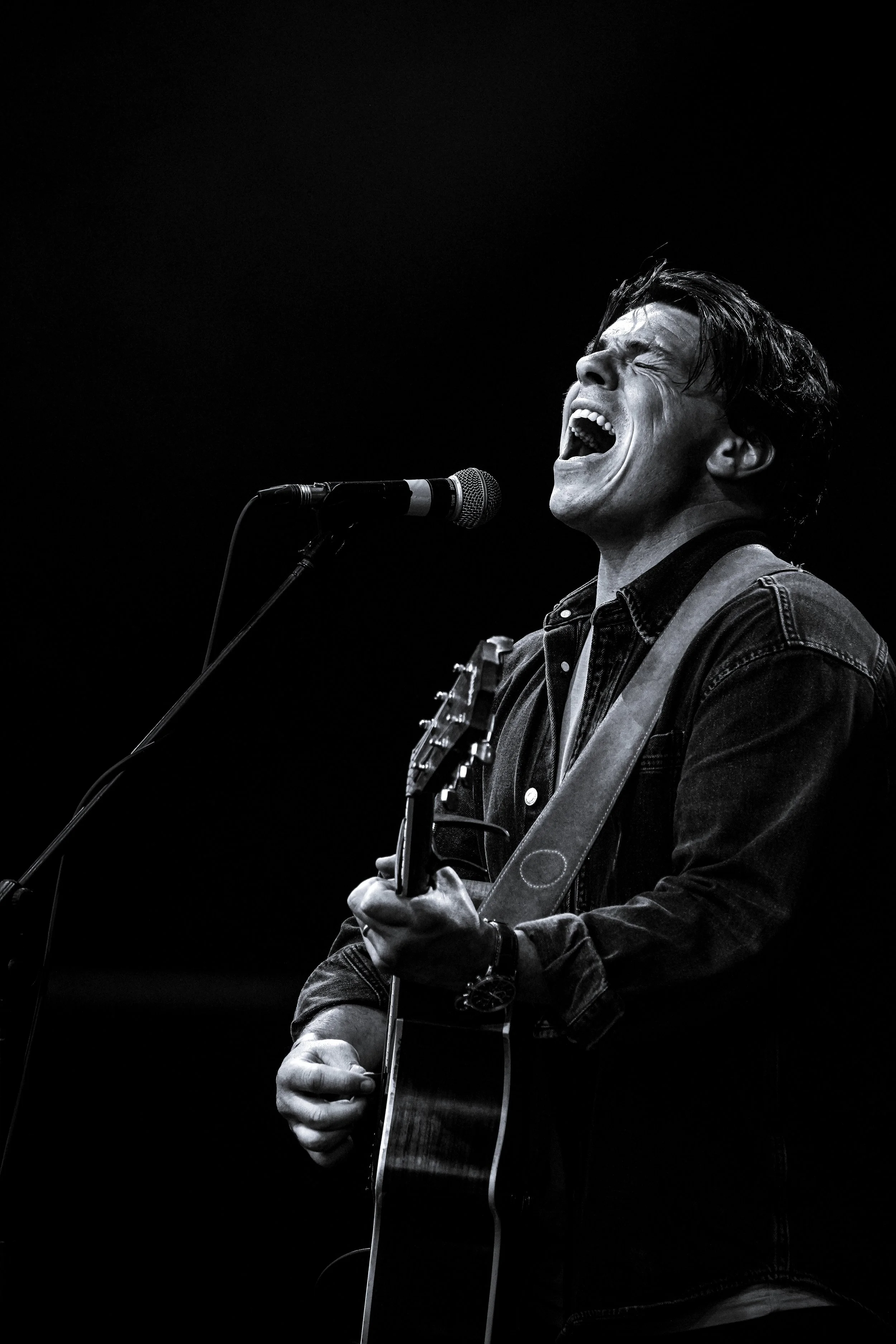 A man passionately singing and playing guitar on stage in black and white.