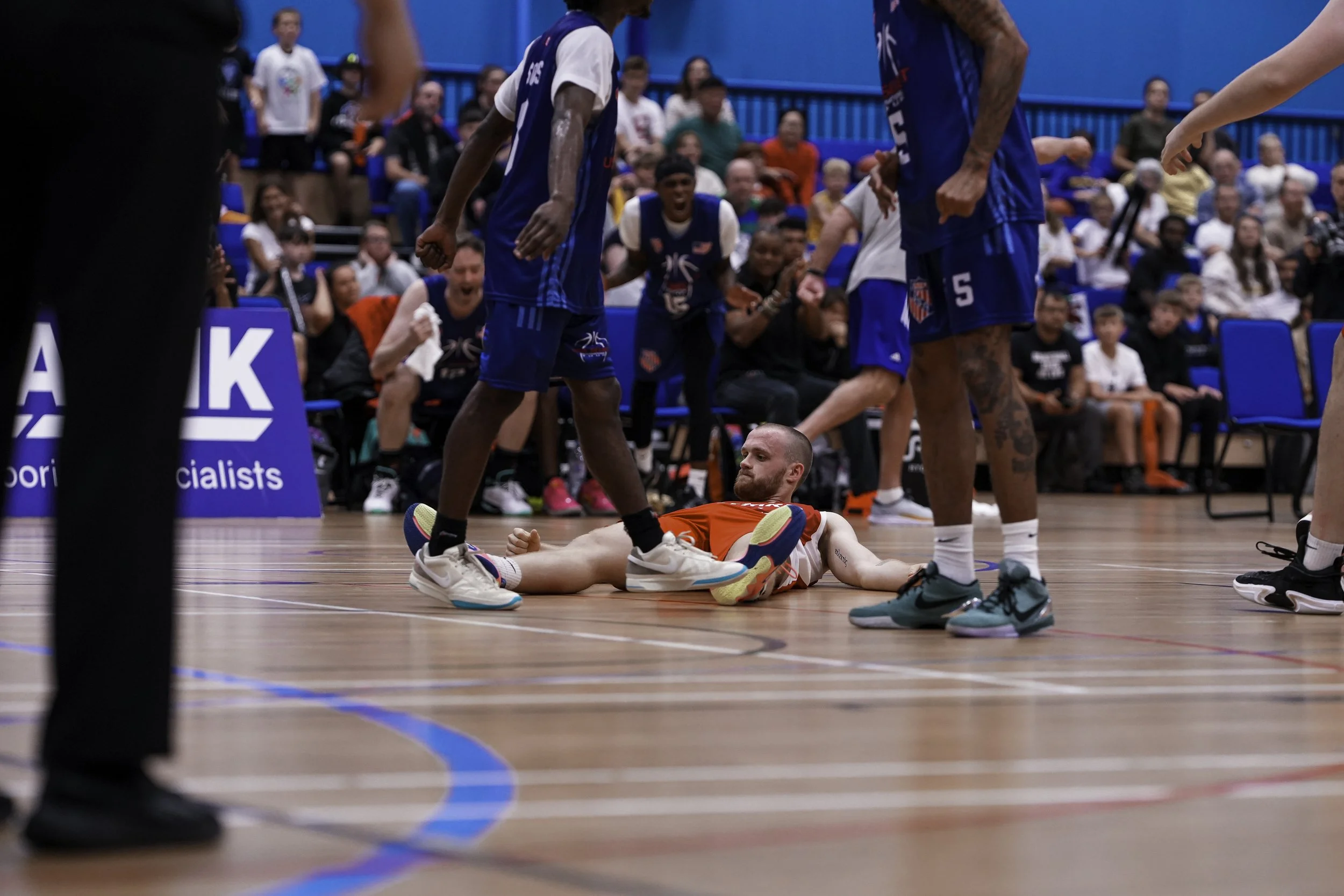 A man is lying on the basketball court floor, surrounded by several players in blue jerseys, with one player standing over him. The scene takes place in an indoor basketball arena with spectators in the background.