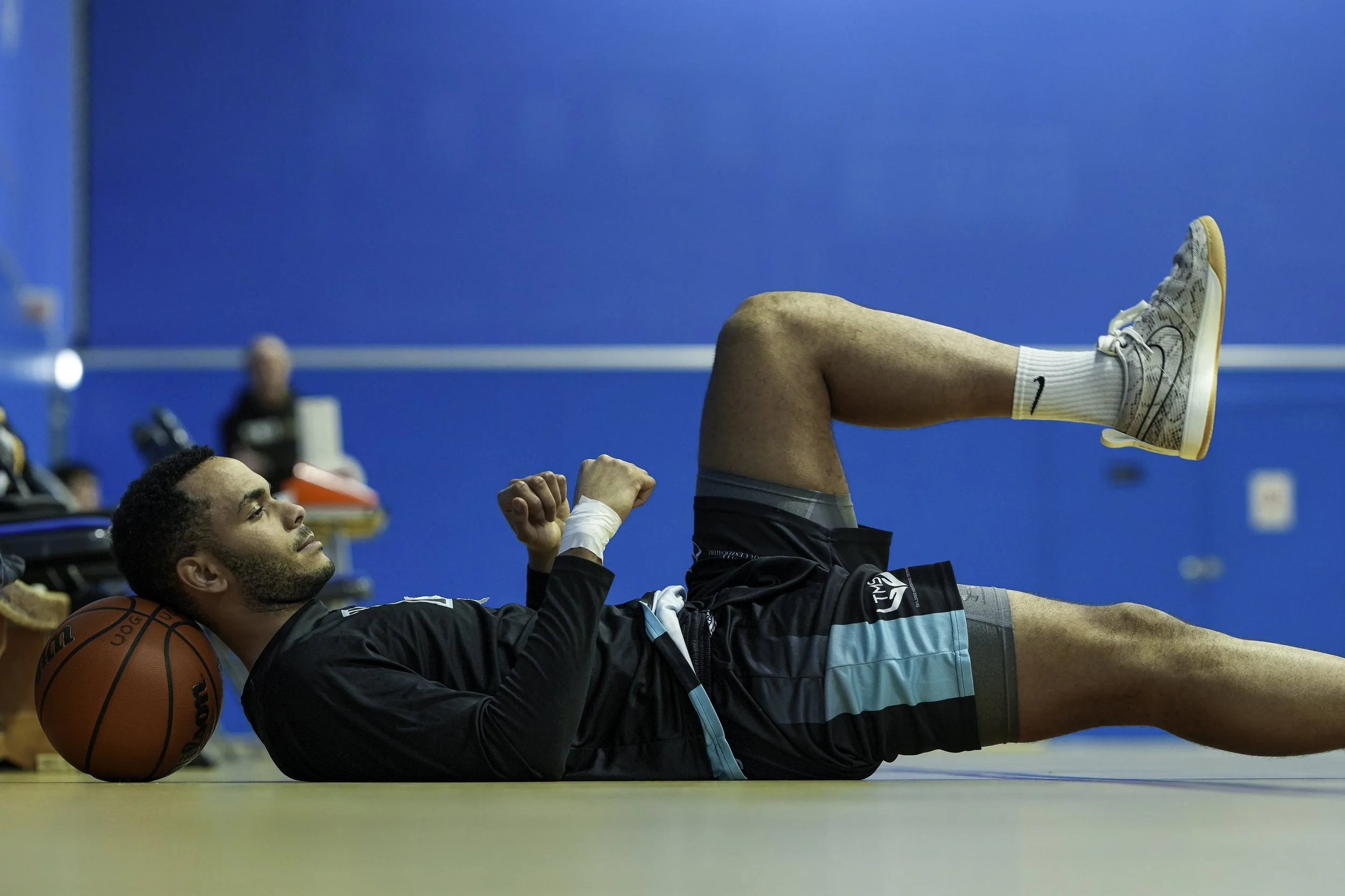 A man lying on the floor with a basketball under his head, stretching on a gymnasium floor, with a blue wall in the background.