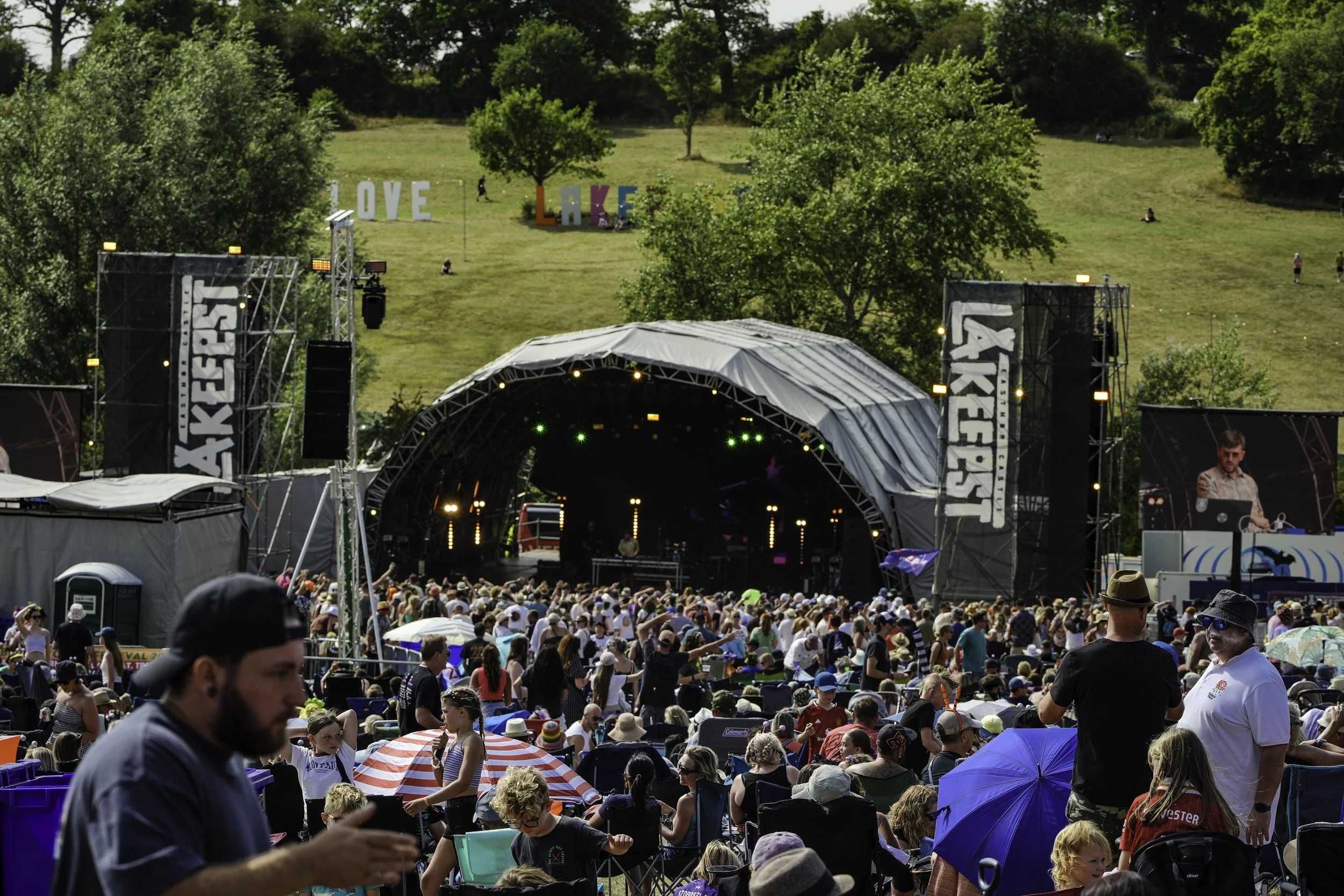 Crowd attending an outdoor concert at a music festival with a large stage, trees, and hillside with colorful 'LOVE LAKE' sign in the background.