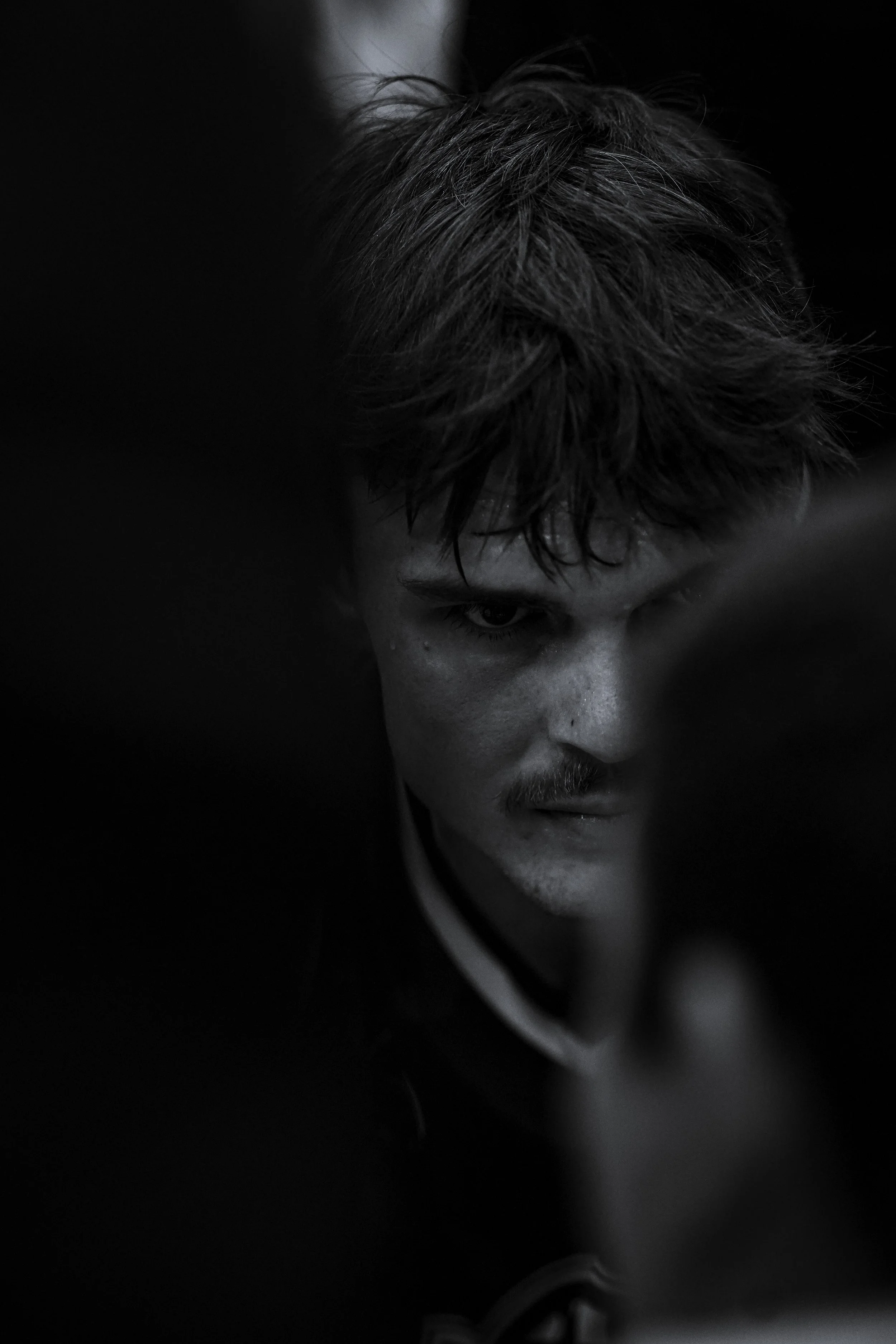 A black and white close-up photo of a young man with wet hair and a mustache, looking intensely at something in front of him, with dark shadows surrounding him.