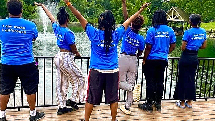Seven people wearing matching blue shirts with the slogan 'I can make a difference' standing on a wooden pier overlooking a lake with a fountain, during daytime.