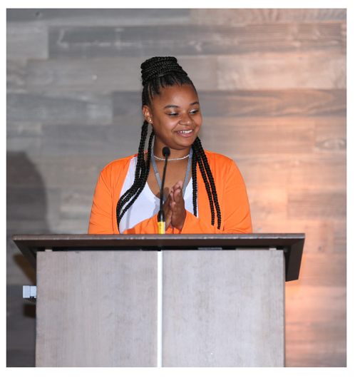 Young woman with braided hair standing behind a podium, smiling, with a microphone, in front of a wooden wall.
