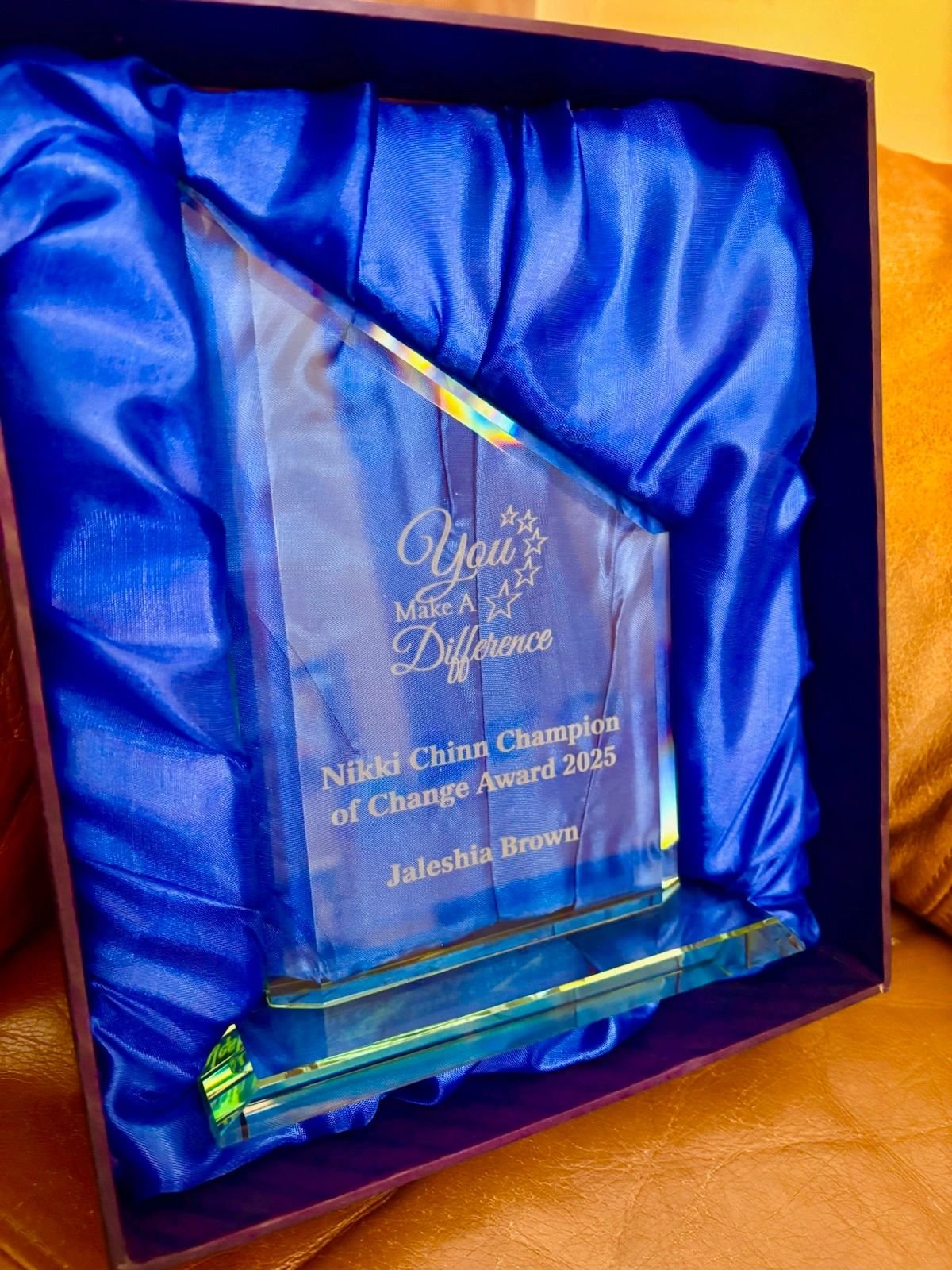 A crystal award with engraved text, presented in a purple box with blue satin lining. The engraving reads: 'You Make A Difference. Nikki Chinn Champion of Change Award 2025. Jaleshia Brown.'