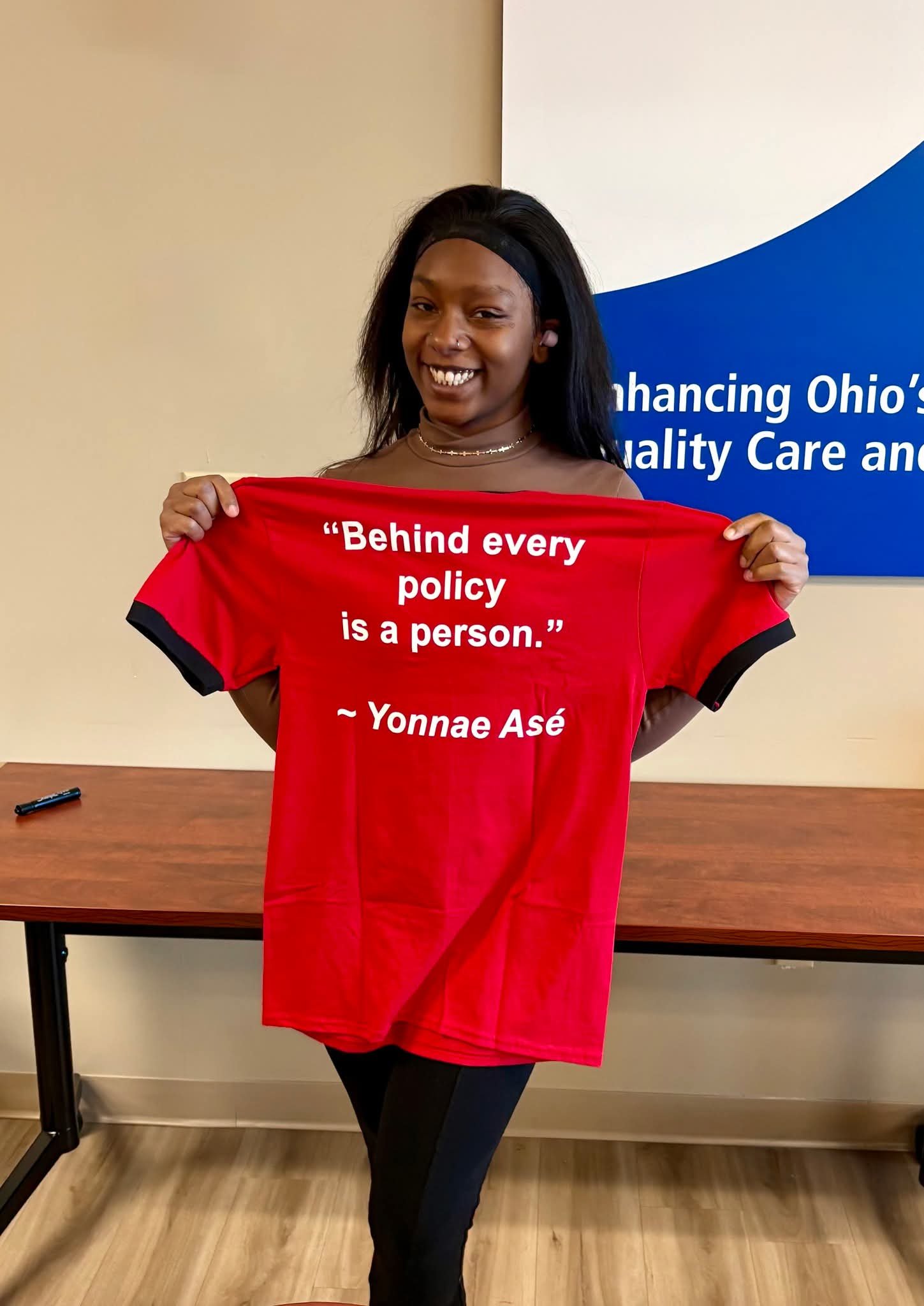 A smiling young woman with dark hair, wearing a headband and dark clothing, holding a red shirt with a quote by Yonnae Asé that says, 'Behind every policy is a person.'