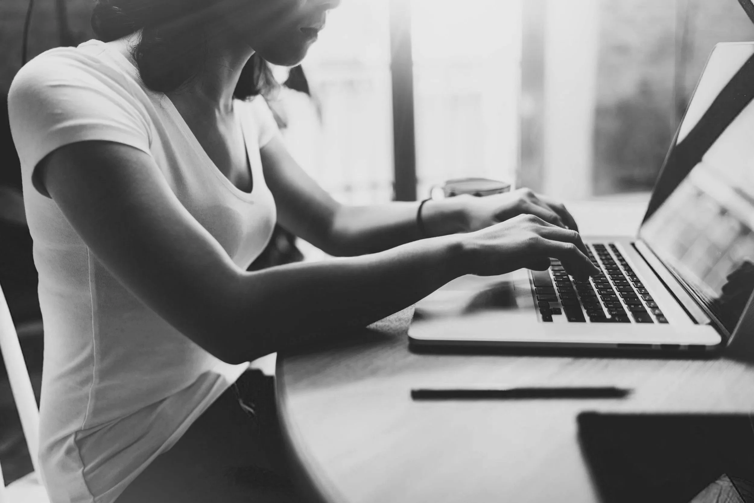 A woman using a laptop at a wooden table near a window, with a cup and a pen nearby.