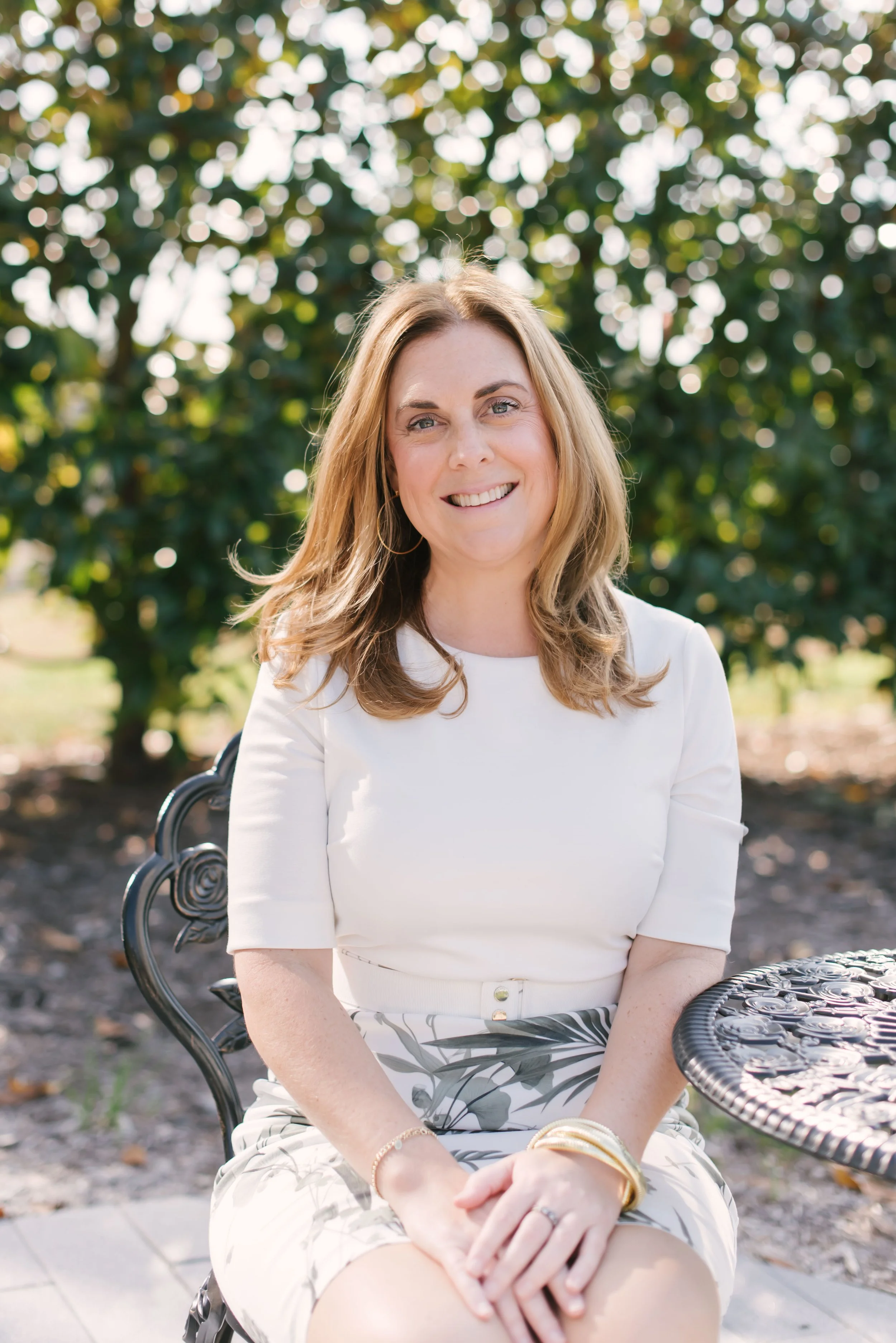 A woman with light brown, wavy hair smiling, sitting outdoors on a wrought iron chair with a decorative table nearby, against a backdrop of green trees and sunlight.