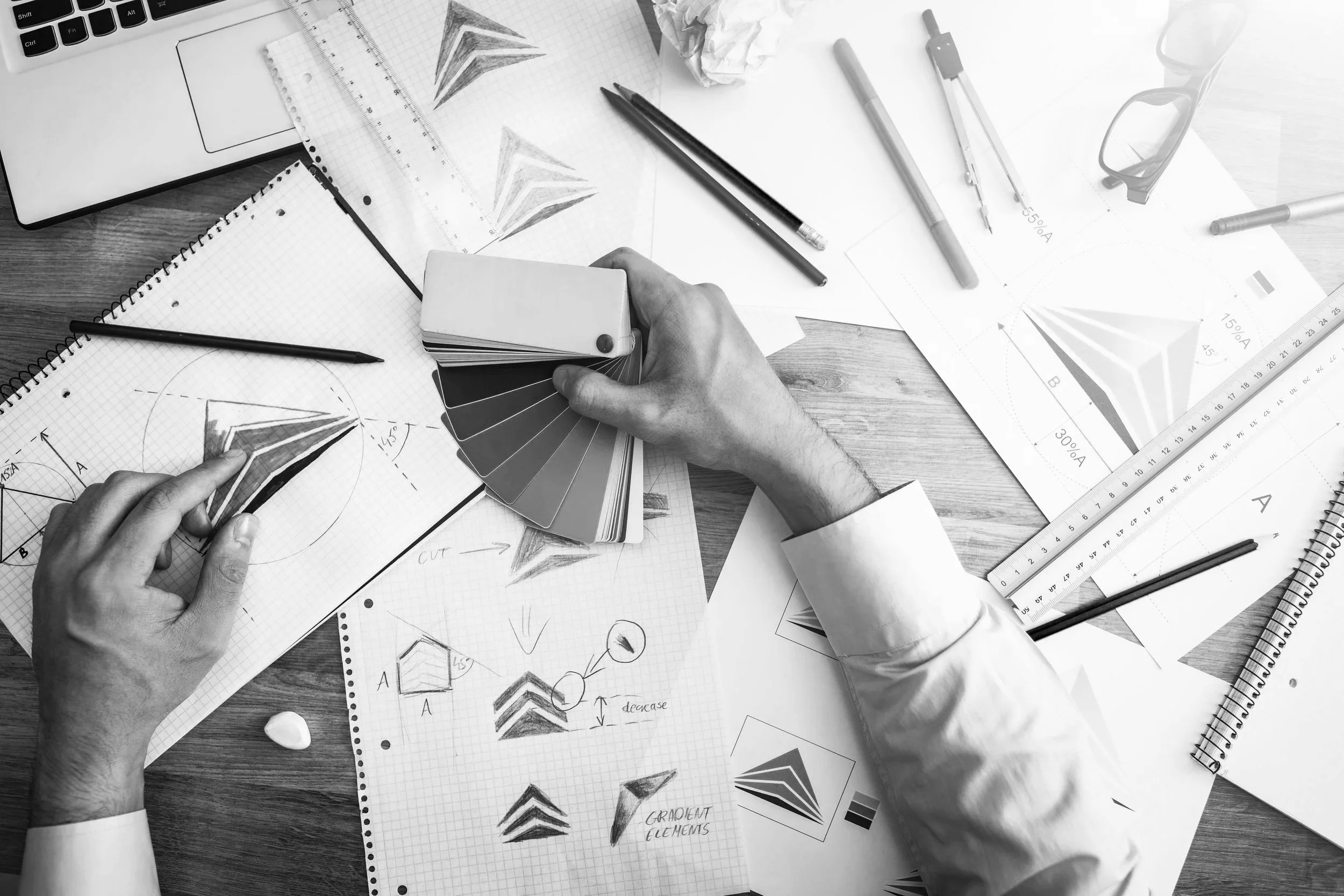 Overhead view of a workspace with design sketches, color swatches, drawing tools, a laptop, and glasses on a wooden desk.