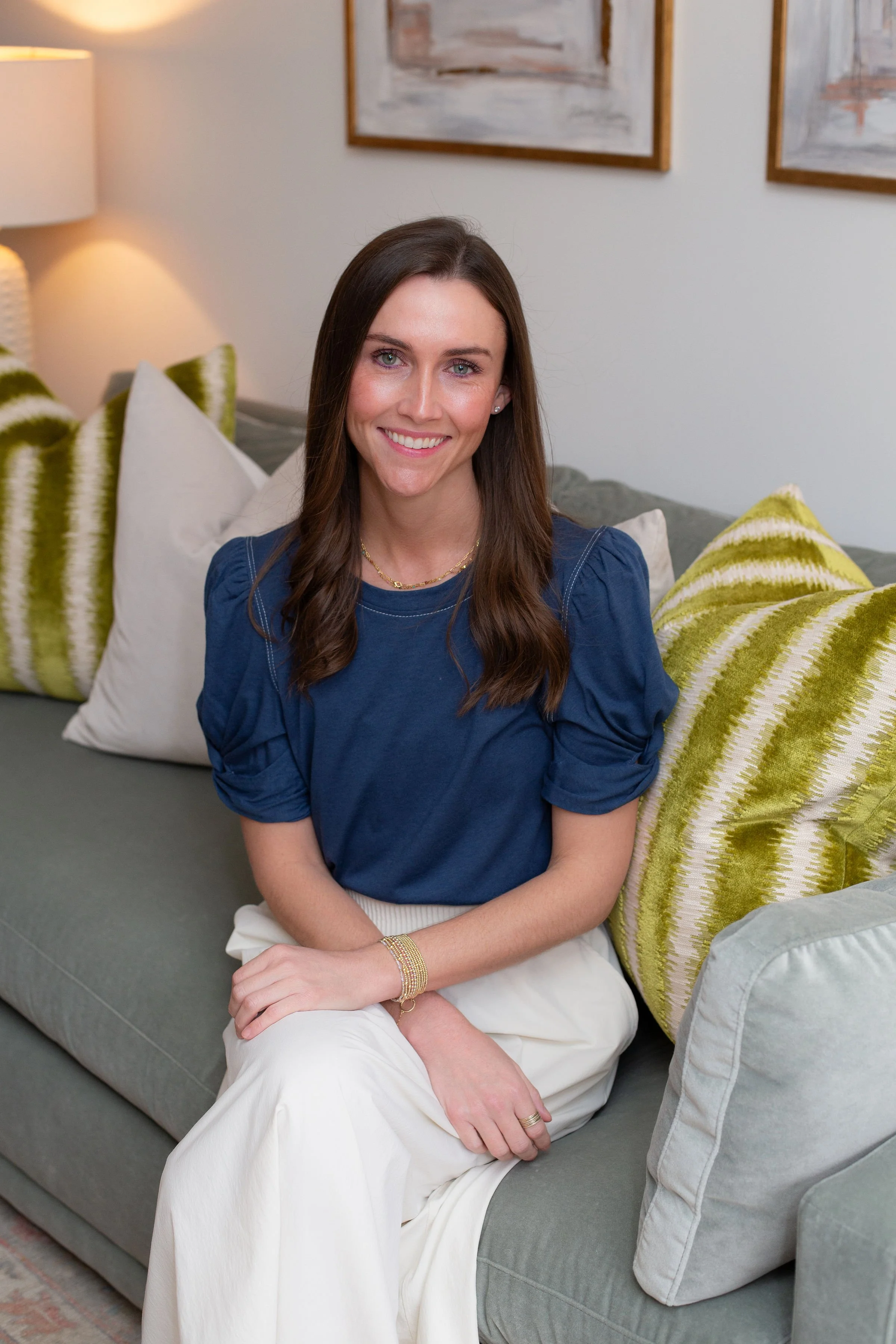 A woman with long brown hair, wearing a blue top and white pants, sitting on a sofa in a living room with a smile.