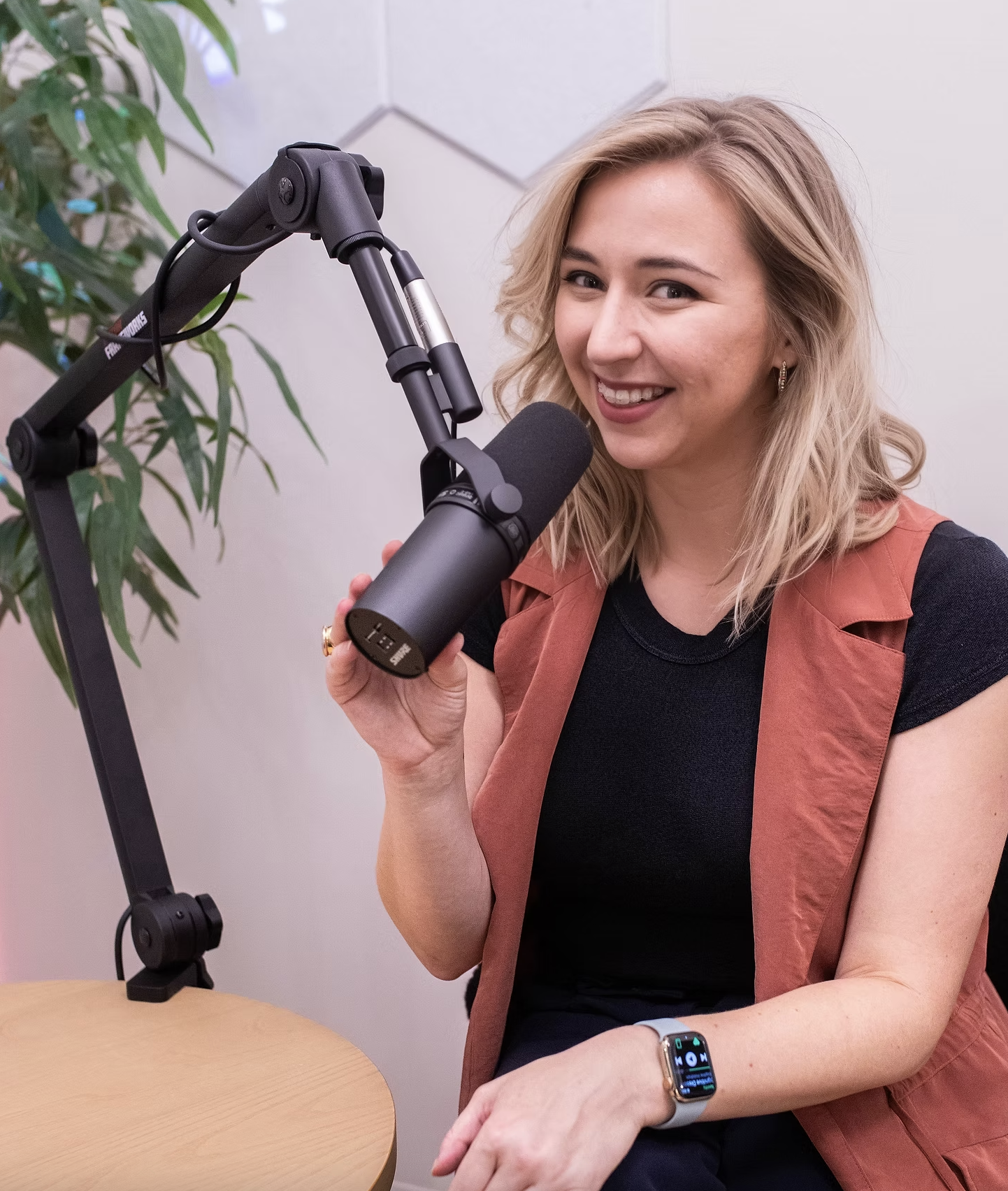 A woman with blonde hair holding a microphone, sitting at a table in front of a microphone arm, wearing a black shirt and an orange vest, with a smartwatch on her wrist, smiling at the camera.